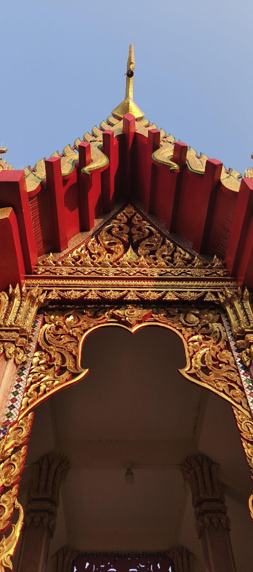 A low-angle shot captures the richly decorated facade of a traditional Thai temple, likely in Bangkok, Thailand, set against a clear blue sky. The upper section of the structure features a prominent golden spire, tapering to a point. Below the spire, the multi-tiered roof is predominantly red, accented with intricate golden and green decorative elements.
Further down, a triangular section of the gable is heavily adorned with elaborate golden bas-relief carvings in a classic Thai 'kanok' style, characterized by flowing, flame-like motifs. This intricate golden ornamentation continues to frame a distinctive archway below. The archway is shaped with a central, stylized curve, outlined in gold and featuring more detailed carvings. Flanking the archway are decorative columns or panels inlaid with small, colorful mosaic tiles in shades of red, blue, green, and gold.
Inside the archway, which leads into a darker interior space, a single light bulb can be seen hanging from the ceiling. The bright, clear blue sky indicates it is daytime, likely morning or afternoon. No people or visible text are present in the image. The scene emphasizes the vibrant colors and detailed craftsmanship typical of Thai sacred architecture.