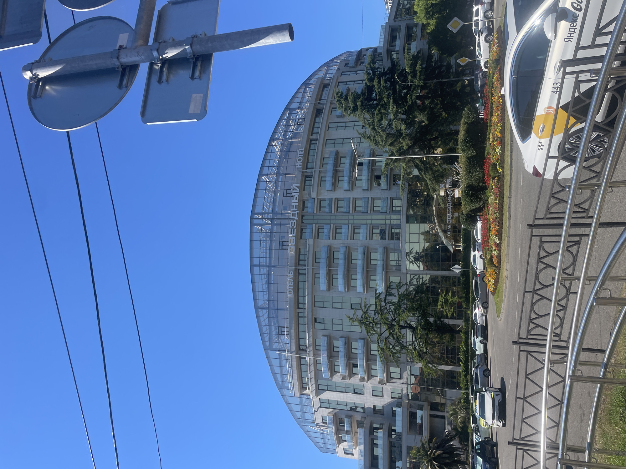 The image shows a daytime exterior view of the Zvezdny Hotel in Sochi, Russia, under a clear blue sky. The hotel is a large, modern building with a distinctive curved, glass-enclosed upper level. The facade features multiple floors with balconies and large windows. In front of the hotel, there is a circular traffic island with colorful flower beds. Several cars are visible on the road, including a white Yandex taxi with the number 443 on its side. There are also traffic signs on a metal pole and overhead power lines. Green trees and palm trees are interspersed around the hotel and the traffic island. The foreground is partially obscured by a metallic railing.