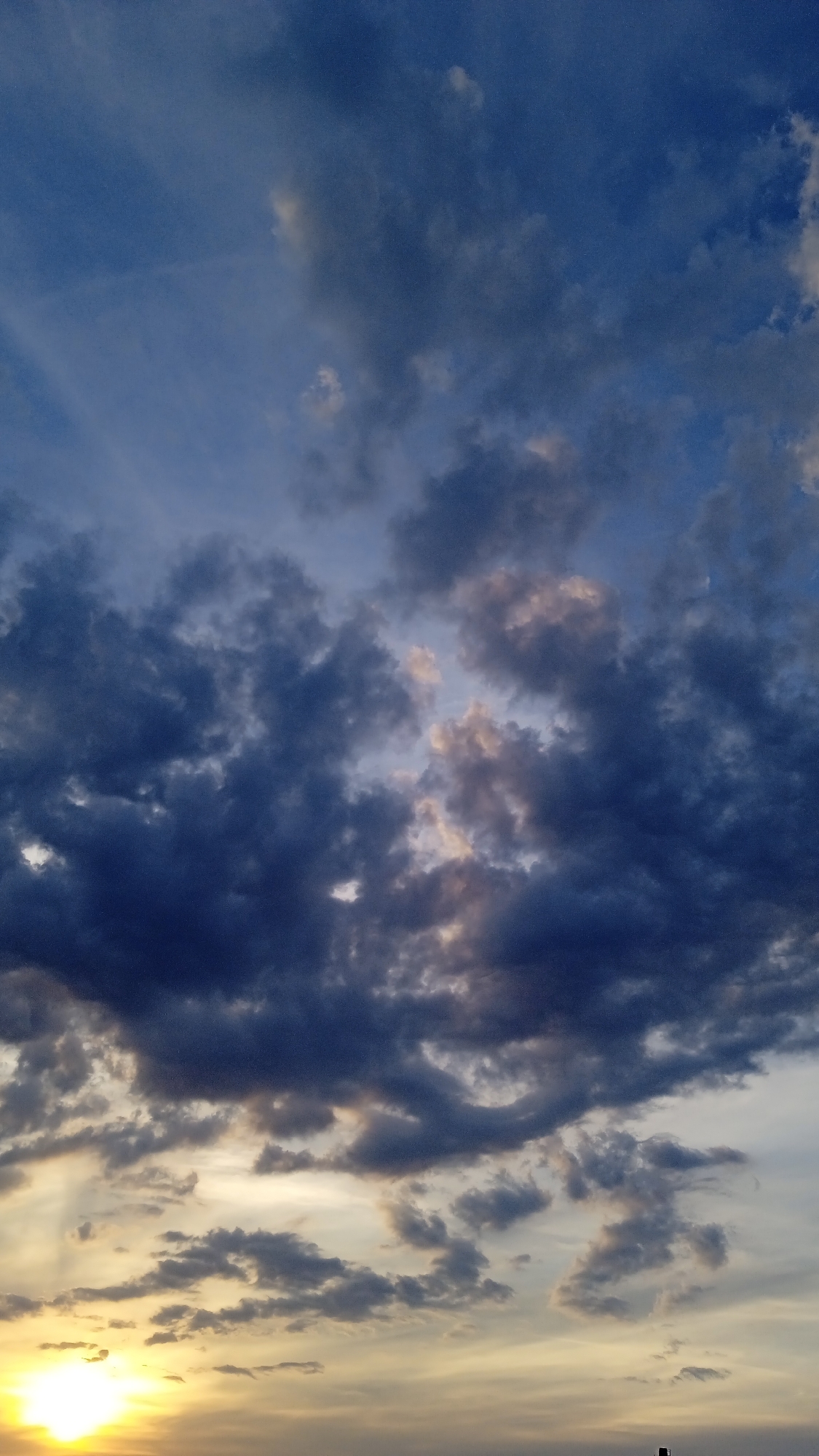 The image shows a dramatic sky at sunset or sunrise. The sky is filled with a variety of clouds, ranging from dark, heavy storm clouds to lighter, wispy cirrus clouds. The sun, a bright yellow orb, is visible in the lower left corner of the frame, casting a warm glow on the clouds around it. The colors in the sky are a mix of deep blues, grays, and oranges, creating a sense of depth and intensity. The clouds are lit from below by the sun, with some parts of the clouds illuminated in shades of pink and gold. The overall impression is one of a powerful and awe-inspiring natural spectacle. There are no people or man-made structures visible in the image, making it a purely atmospheric scene.