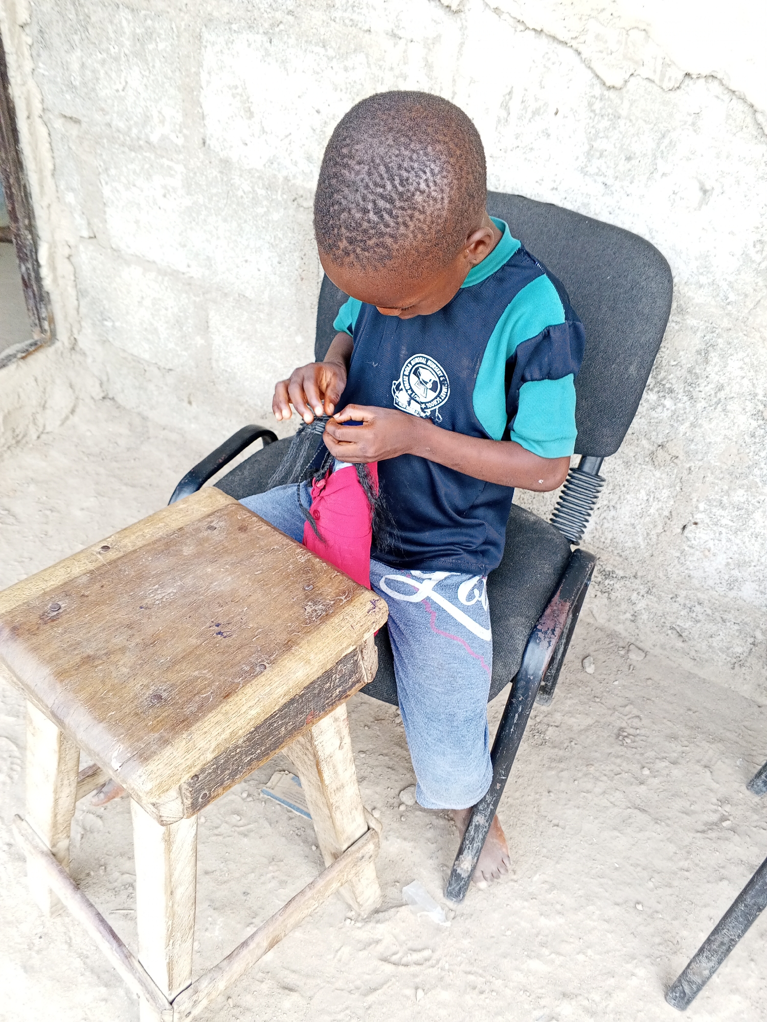 A young child sits in a black office chair positioned in front of a small, wooden table in Unknown, Unknown. The child, wearing a blue and teal shirt and blue jeans, appears to be manipulating a black hair extension. The environment is simple, with a roughly textured wall behind the child and a sandy surface underneath the chair and table.
