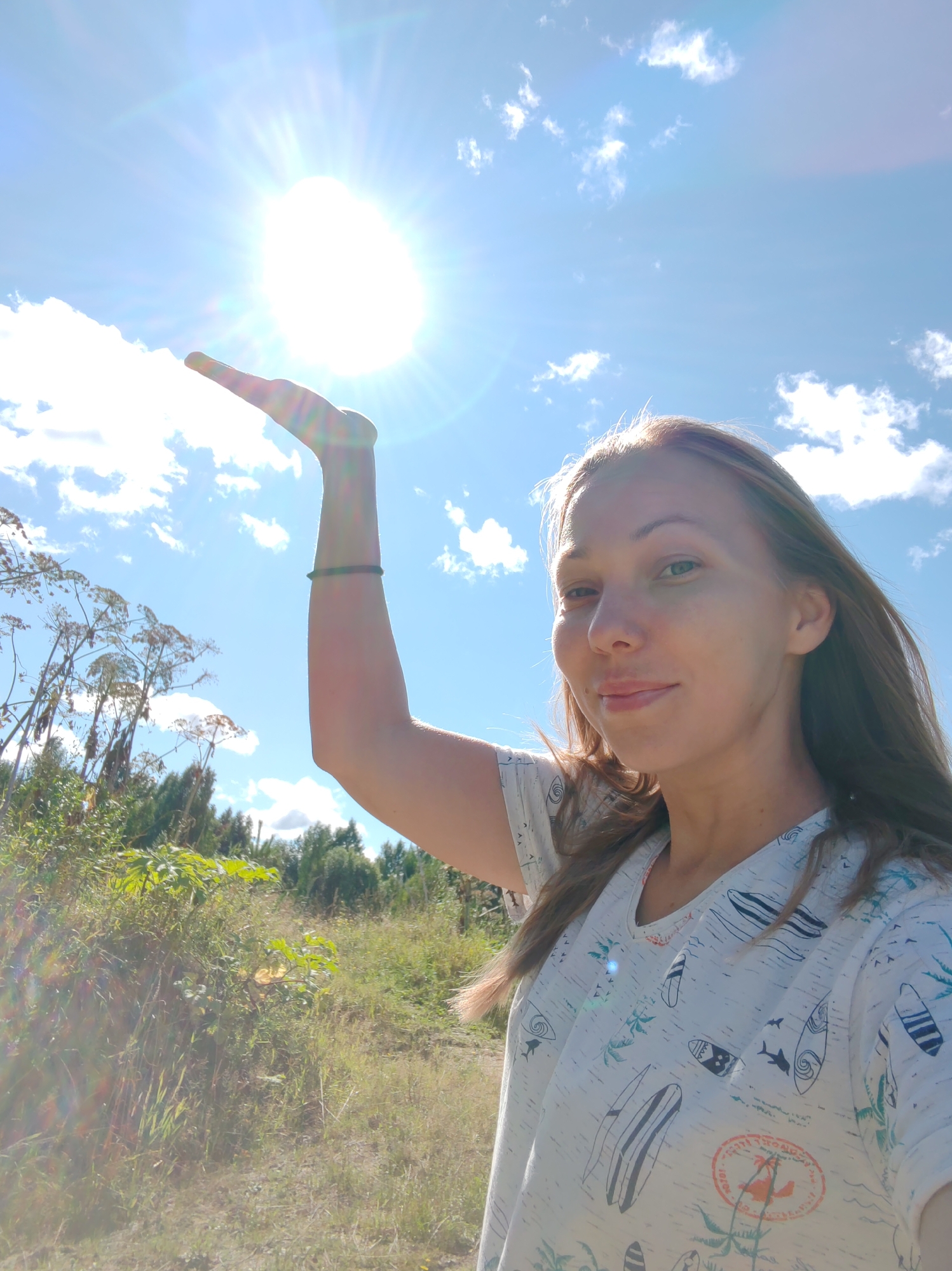 A woman is taking a selfie outdoors. She has long, light brown hair and is wearing a white t-shirt with a surf-themed pattern. Her left arm is raised, and her palm is extended upwards as if she is holding the sun. The sun is very bright and prominently featured in the upper part of the image, with visible sun flares. The background consists of a clear blue sky with scattered white clouds, green vegetation, and trees. The location is identified as Чухлэм, Russia.