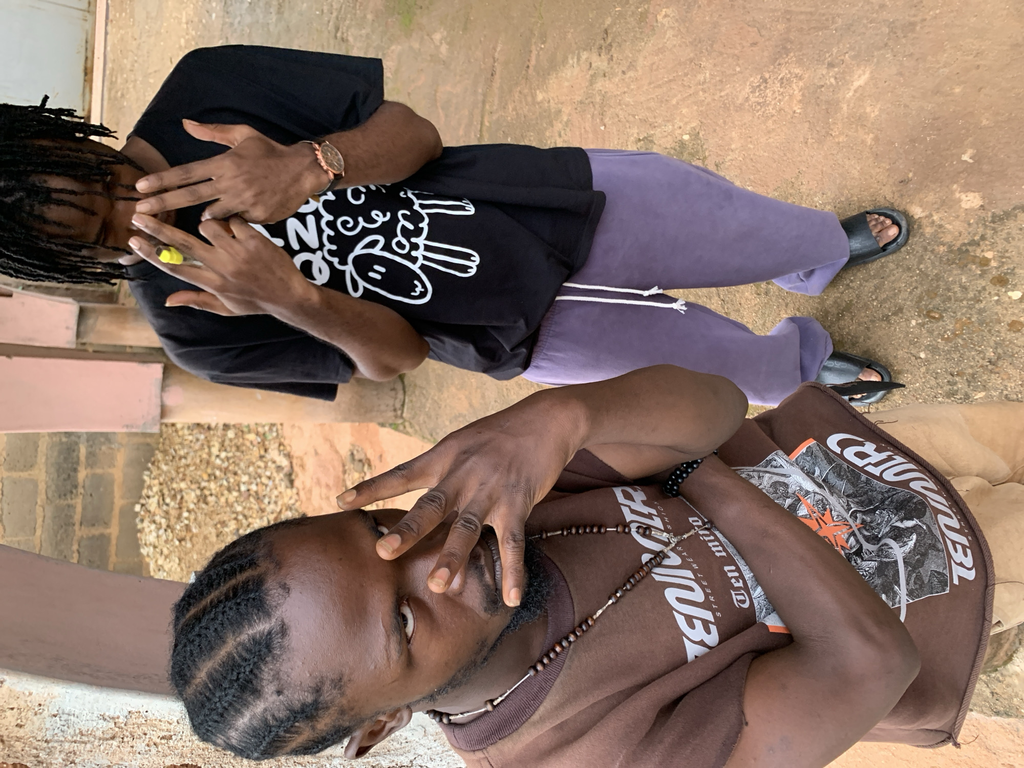 Two young Black males, appearing to be in their late teens or early twenties, are positioned in an outdoor, urban environment. The male in the foreground, on the left, has braided hair and a goatee, and is wearing a brown t-shirt with a graphic print and a beaded necklace. He is making a hand gesture with his right hand, fingers extended, with his left arm crossed over his chest. His gaze is directed towards the camera with an intense expression. The second male, on the right, has dreadlocks and is wearing a black t-shirt with a white sheep graphic, and light purple sweatpants. He is holding a yellow object, possibly a pen or marker, and has both hands raised with fingers extended in a gesture. Both individuals are wearing casual footwear. The background consists of a rough, textured wall to the left, and a reddish-brown, uneven ground surface. In the distance, there are piles of rubble and what appears to be a building constructed from brick and possibly plastered walls, suggesting a developing or semi-developed area. The scene is lit by natural daylight, casting soft shadows. The overall impression is one of youthful engagement, potentially in a street or neighborhood setting within Jos, Nigeria, given the architectural and environmental cues.
