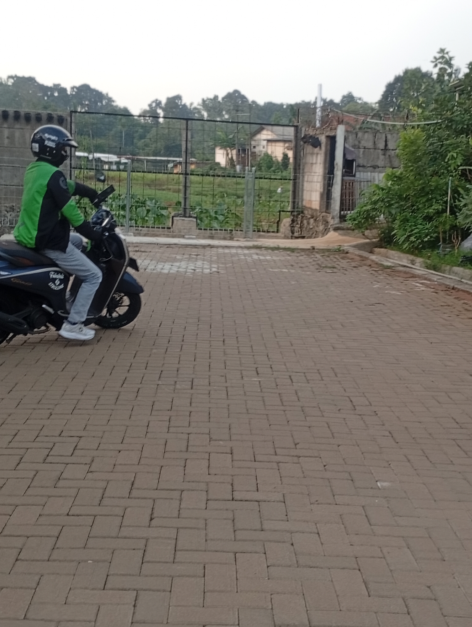 A person wearing a black helmet with a green and black jacket rides a dark-colored scooter on a brick-paved road. The scooter has a logo on its side. The background features a metal fence, trees, and some buildings. The scene appears to be outdoors in Kemang, Indonesia.