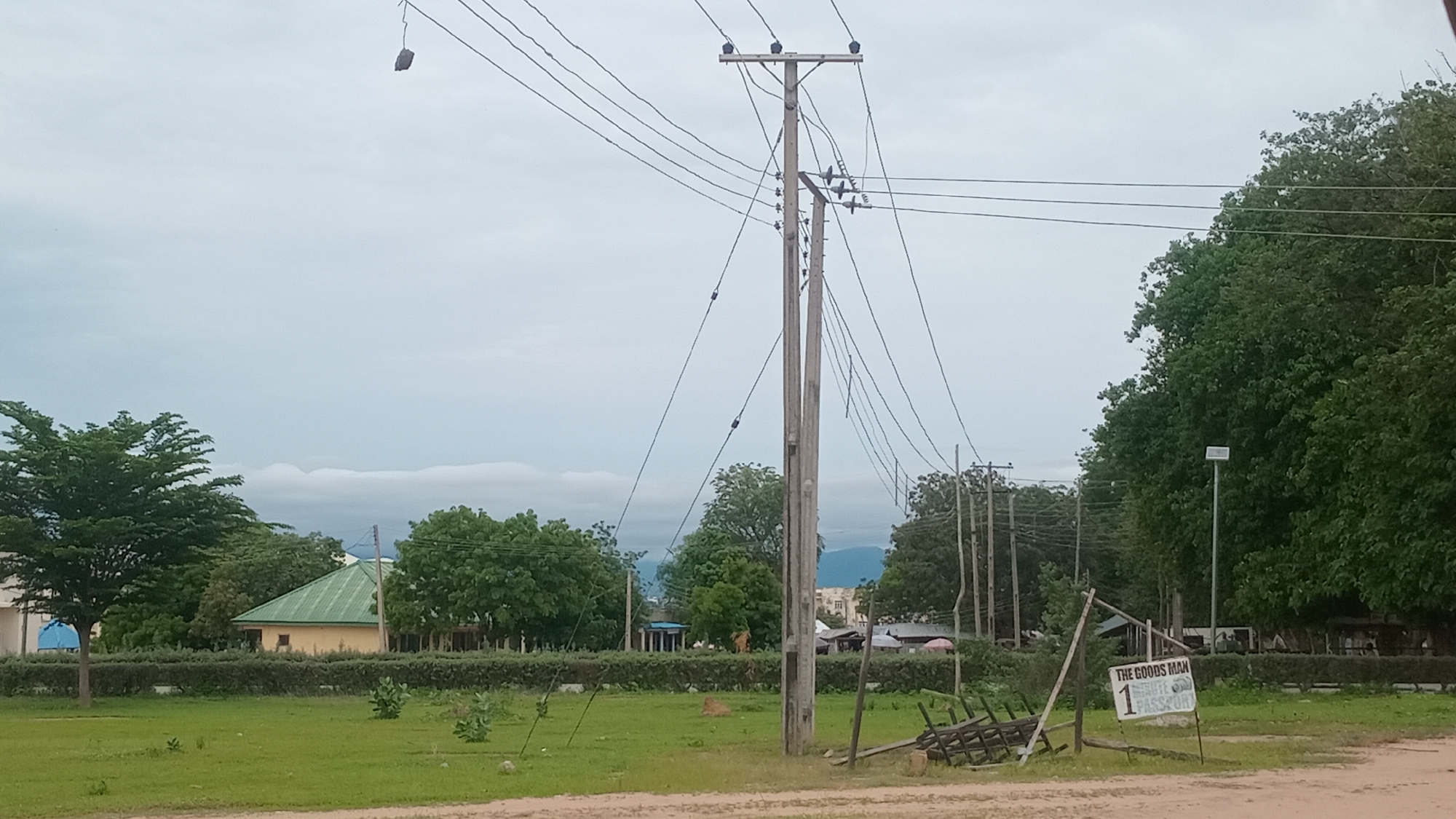 This is a medium-distance, eye-level, outdoor, daytime photograph of a scene in Jalingo, Nigeria. The sky is overcast and filled with gray clouds. In the foreground, a dirt road is visible. To the right of the road, a wooden sign reads "THE GOODS MAN ROUTE 1 PASSPORT". There is a small stack of metal objects resembling a dismantled cart or sled behind the sign. A large wooden utility pole with numerous electrical wires stretches across the frame, with a secondary, shorter pole to its right.
In the middle ground, a neat green lawn is bordered by a low, manicured hedge. Behind the hedge, a light yellow building with a green, sloped roof is visible, flanked by trees. More trees and a few buildings are visible in the background, with distant hills under the cloudy sky. The overall impression is of an area that is partly developed with infrastructure and greenery.