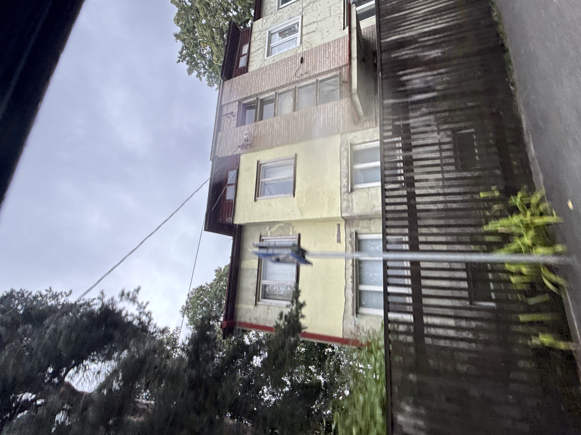 The image shows a multi-story building with a light yellow facade and some brickwork, likely in Tallinn, Estonia. The building has several windows, some with white frames and others with blinds visible. There is a red-colored trim on the upper part of the building and also a red downspout on the left side. A dark wooden fence runs across the foreground. A tall metal pole with a blue sign is positioned in front of the fence and the building. Trees are visible on the left and right sides of the building, with the sky above appearing overcast. The overall scene depicts an exterior view of residential architecture under a cloudy sky.