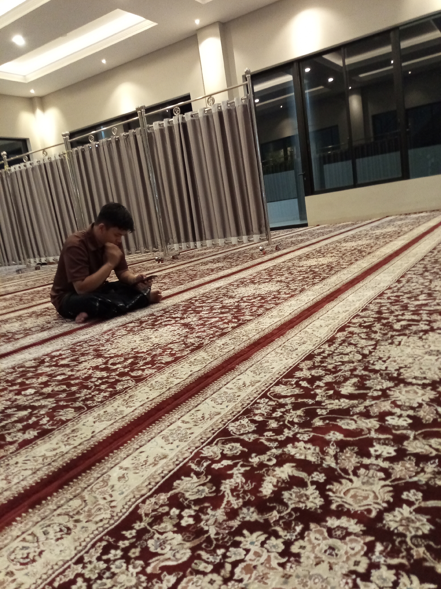 A man is seated on a carpeted floor, looking down at a mobile phone. He is wearing a brown shirt and dark pants. The room appears to be a prayer room, with a large, ornate carpet covering the floor. Long, grey curtains hang along one wall, and large windows with dark frames are visible on another wall. The lighting suggests it is indoors, possibly at night or in a dimly lit space, with recessed ceiling lights illuminating the room. The overall impression is one of quiet contemplation or personal time. The location is identified as Cihanjuang, Indonesia.