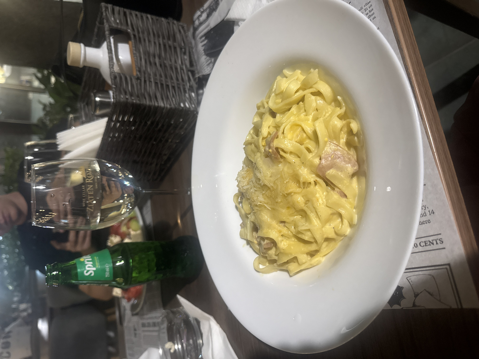 The image depicts a dining table scene at the Zlaten Roz Wine Cellar in Petrich, Bulgaria, as indicated by the text "WINE CELLAR ZLATEN ROZ SINCE 2010" etched on the wine glass. In the foreground, a large white ceramic plate holds a serving of creamy, yellow tagliatelle pasta, studded with pieces of bacon or pancetta and topped with grated cheese. To the left of the pasta plate, a 250ml green glass bottle of Sprite stands upright. Adjacent to the Sprite, a stemmed wine glass, partially filled with white wine, is prominently displayed. To the right of the wine glass, a dark woven table caddy contains folded white paper napkins, a small cylindrical metal container, and a white ceramic bottle with a light brown stopper, likely for oil or vinegar. Further in the background, a blurry individual, appearing to be a young adult male, is seated at the table, partially obscured and holding a mobile phone. A small plate with a salad containing red and white elements is visible behind the Sprite bottle. The table surface is dark wood, and the overall environment is an indoor restaurant with dim, ambient lighting.