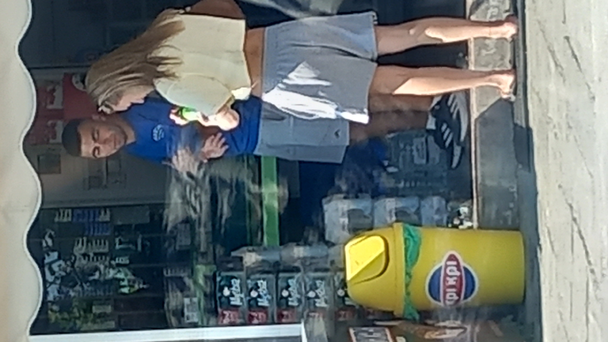 The image shows two people, a man and a woman, standing in front of what appears to be a small convenience store or kiosk. The man is wearing a blue t-shirt and grey shorts. The woman is wearing a light yellow crop top and grey shorts, and she is holding a green bottle. A yellow trash can with a blue and red logo is to the left of the people, and the logo on the trash can reads "ΕΦΗΜΕΡΙΔΕΣ" and "ΤΥΠΟΣ", which are Greek words for "newspapers" and "press" respectively, often seen on newsstands and kiosks in Greece. Products are displayed on shelves behind them, visible through a glass or transparent surface. The ground in front of the store is paved. The overall setting suggests a street-level establishment in Greece, likely in a town or city. Given the presence of Greek text and the style of the kiosk, it is highly probable that this scene is located in the city of Kos, Greece.