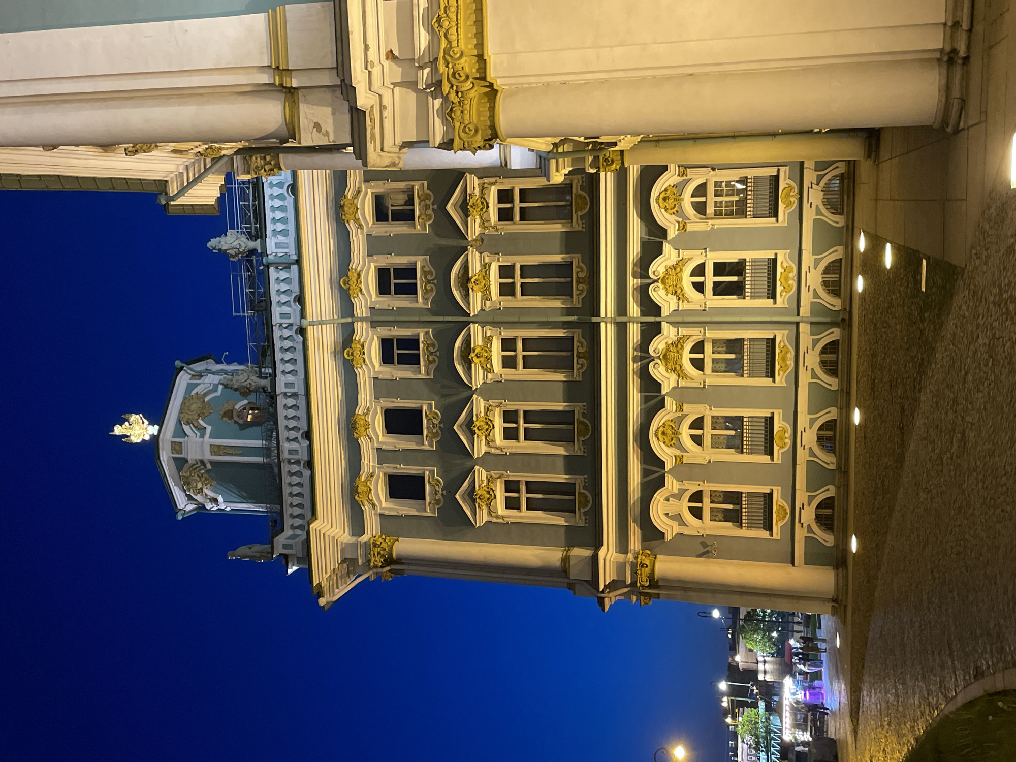 This image captures a grand, ornately decorated building at night in Saint Petersburg, Russia. The sky is a deep, clear blue, characteristic of twilight or early evening, creating a stark contrast with the brightly illuminated architecture.
The primary subject is a multi-story building rendered in a rich Baroque or Rococo style. Its facade is a distinctive pale blue-grey, complemented by crisp white architectural trim and abundant golden ornamentation. The building features numerous large windows of various shapes – rectangular, arched, and semi-circular – many framed by elaborate golden decorative elements such as pilasters, cornices, and pediments. Intricate golden sculptures, relief work, and decorative capitals adorn columns and other structural elements, showcasing detailed craftsmanship. On the left side, a portion of a small dome or cupola is visible, topped with golden finials and sculptures, hinting at a palace church or a significant part of a larger complex. Gargoyle-like figures or decorative heads are also discernible on some cornices and columns.
In the lower right section of the image, a paved area, likely a street or courtyard, is illuminated by ground lights and more distant streetlamps. A few indistinct figures of people are visible, suggesting some nocturnal activity, possibly near an outdoor stall or establishment that is also brightly lit.
The overall scene evokes a sense of historical majesty and opulence, typical of Saint Petersburg's imperial architecture. The artificial lighting dramatically emphasizes the building's intricate details and grandeur against the dark night sky. No discernible text is visible within the image.