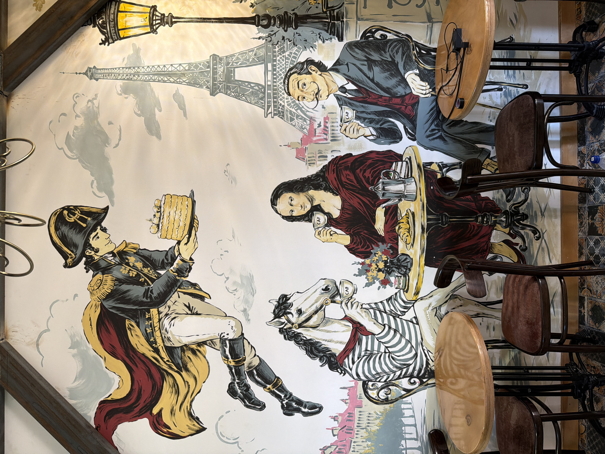 The image is a mural painted on a wall, likely in a cafe. The mural depicts a surreal scene set against a Parisian backdrop, featuring the Eiffel Tower and Haussmannian buildings.
In the upper portion of the mural, a stylized character resembling Napoleon Bonaparte in military attire, complete with a cape and bicorne hat, is depicted floating and holding a cake.
In the lower portion, three figures are seated at tables. To the left, a white horse with a human-like torso is dressed in a striped shirt and red scarf, holding a cup of coffee. Next to it is a table with a croissant, cake, and a vase of flowers. In the center sits a figure resembling the Mona Lisa, also holding a cup of coffee. To the right is a figure with a prominent mustache, reminiscent of Salvador Dalí, dressed in a suit and vest, also holding a cup of coffee.
The cafe itself appears to have tables and chairs in the foreground, with wooden tables and dark chairs. The flooring has a tiled pattern. The lighting in the real-world setting suggests a warm, ambient light, likely from lamps and possibly natural light filtering in.
While the mural's theme is Parisian, the image was taken at a cafe located in Tashkent, Uzbekistan. The specific cafe is likely "Café 1963" located at 38 Amir Temur Street, Tashkent, Uzbekistan, given the popular use of this mural in photos from that establishment.
