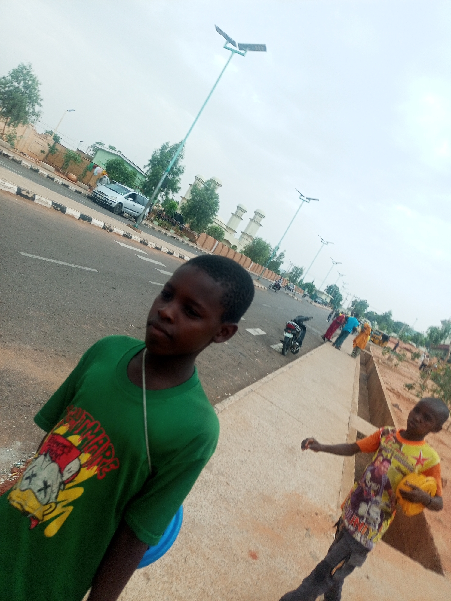 The image depicts two boys on a sidewalk in Gombe, Nigeria. One boy, wearing a green t-shirt, is in the foreground, while the other, wearing an orange and yellow shirt and holding a yellow object, is slightly behind him. In the background, there is a road with a silver car and a motorcycle, as well as several other people walking. The architecture of a building with white turrets and several light poles are visible. A freshly planted area is visible to the right of the sidewalk. The sky is overcast.