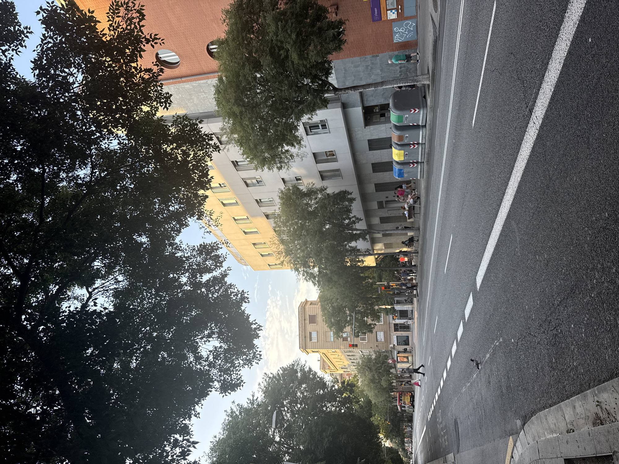 The image depicts a street scene in Barcelona, Spain. The perspective is from street level, looking down a wide avenue lined with trees and buildings. The sky is visible through the dense canopy of leaves from mature trees that dominate the foreground and midground.
In the midground, several multi-story buildings are visible. To the right, a prominent brick building with circular windows is noticeable. Adjacent to it, a lighter-colored building with grey facade panels and numerous windows with shutters stands. Further down the street, other buildings with various architectural styles and colors, including yellow and beige, can be seen.
The street itself is paved and appears to be a busy thoroughfare with multiple lanes. White lane markings and a dashed white line demarcate the traffic lanes. Several cars and a yellow bus are present on the road. Pedestrians are also visible: a man is crossing the street at a pedestrian crossing, and a small group of people are seated on a bench near the sidewalk, while others are walking along the sidewalk.
In the foreground, the sidewalk and curb are visible. There are several large, colorful recycling bins (blue, yellow, brown, and green) lined up along the sidewalk in front of the buildings on the right. A pigeon is standing on the road in the lower left quadrant of the image.
The lighting suggests it is late afternoon or early evening, with the sun casting long shadows and illuminating the upper portions of the buildings. The overall atmosphere is that of a typical urban environment in a European city, with a mix of residential and commercial structures.