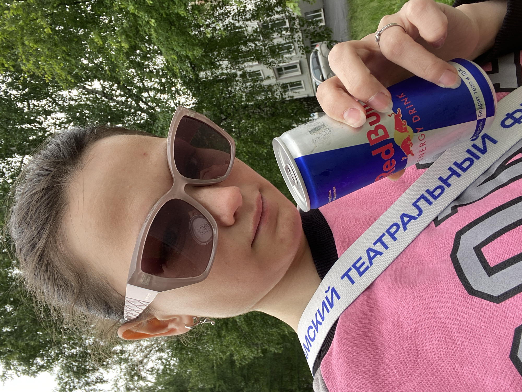 The media file depicts a person, likely female, shown from the chest up in an outdoor setting in Chaikovsky, Russia. The image is oriented with the subject's head significantly tilted to the left, almost 90 degrees. The person wears large, light-colored framed sunglasses, which obscure their eyes, and a pink long-sleeved top with a black trim at the collar.
The subject holds a silver and blue Red Bull energy drink can in their right hand, with a silver ring visible on a finger of their left hand (which is also holding the can). The Red Bull logo and "ENERGY DRINK" are prominent on the can. Visible Cyrillic text on the can reads "Таурин, Бодрит тело и дух." (Taurine, Invigorates body and spirit.) and a partially visible warning label that includes the word "треблять" (to consume), indicating a health advisory. The word "eternal" is visible on the temple of the sunglasses.
A light-colored strap with blue Cyrillic lettering crosses diagonally over the person's chest. The visible text on the strap is "МСКИЙ ТЕАТРАЛЬНЫЙ", which likely refers to a theatrical entity (e.g., "Permskiy Teatralny" - Perm Theatrical). The person has dark hair, and small hoop earrings are visible on their left ear.
The background is an out-of-focus outdoor scene, dominated by green tree foliage. A white multi-story building with windows and a partially visible grey car can be seen through the trees, indicating an urban or suburban environment. The lighting suggests it is daytime, possibly overcast. The subject's facial expression, with sunglasses covering their eyes, appears neutral.
