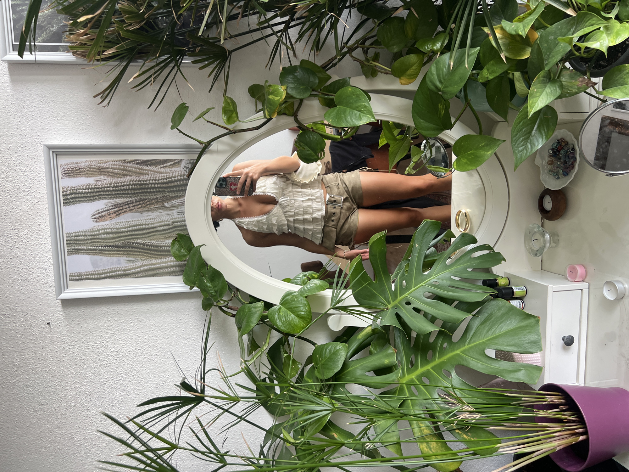 This is a vertical, eye-level, medium shot of a woman taking a mirror selfie. The setting appears to be a bright, indoor space, possibly a bedroom or dressing area, with several large potted plants framing the shot.
The woman, reflected in an oval mirror with a white frame, is wearing a ruffled white top and tan shorts. She is holding a smartphone in her right hand to take the photo. Her left arm is bent at the elbow and resting on a surface. The mirror is positioned above a white dresser or vanity that has a small drawer with a dark knob. On the dresser are several small objects, including a clear dish with colorful beads, a small wooden holder with a candle, a clear glass votive holder, and a pink and a white votive candle. There are also several small bottles, likely essential oils, arranged on the top of the dresser.
To the left of the mirror, a framed picture of cacti hangs on a textured white wall. Lush green foliage from various plants fills the foreground and surrounds the mirror and dresser. A large monstera leaf is prominent on the right side of the frame, and a papyrus plant is leaning forward from a purple pot on the bottom right.
The lighting suggests it is daytime. There are no visible emotions conveyed by the woman as her face is partially obscured by the phone. No text is visible in the image. The presence of indoor plants and home decor suggests a domestic setting.