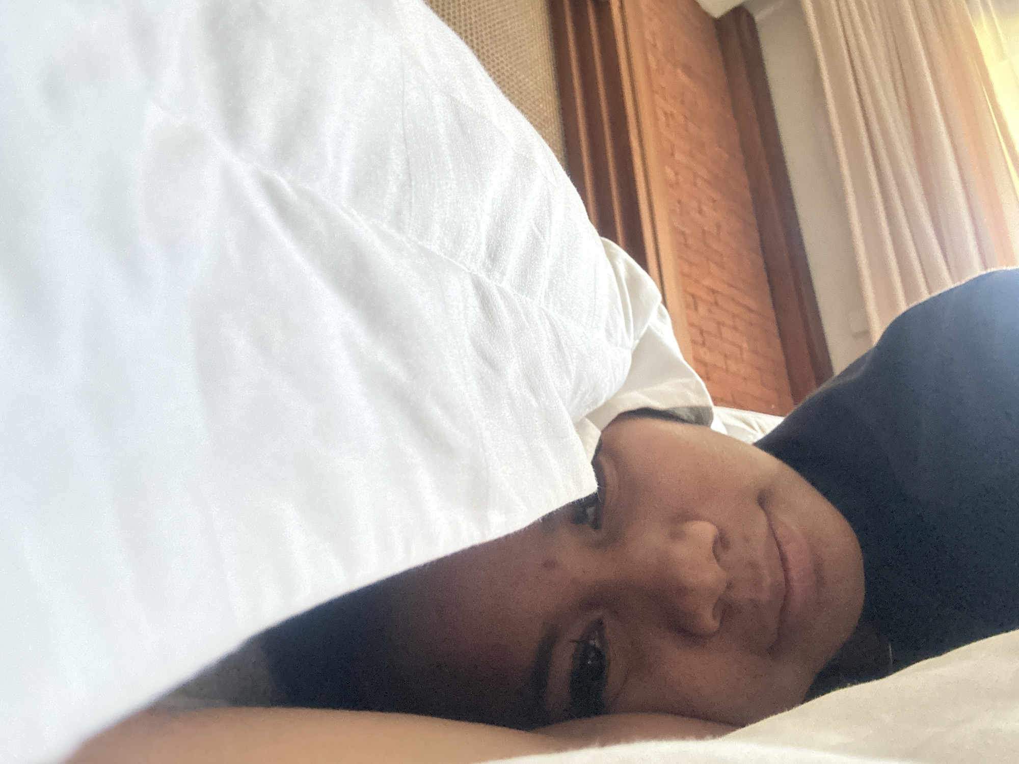 A close-up, eye-level shot depicts a person lying in bed, partially covered by a white blanket, presumably in Ubud District, Indonesia. The individual, with medium skin tone and dark hair, is wearing a dark-colored garment. Their head rests on a white pillow or sheet. Part of the face is visible, showing a relaxed expression. The background features a wall with vertical wooden panels and white curtains.
