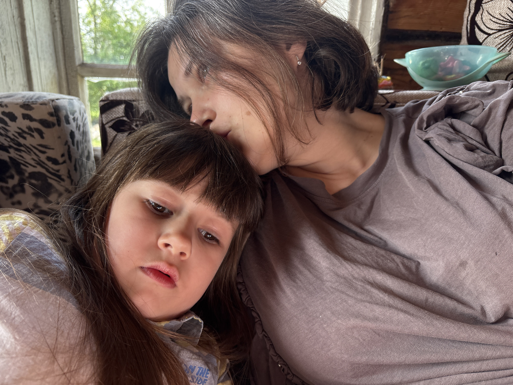 A woman with short brown hair is kissing the forehead of a young girl with long brown hair. The woman is wearing a purple-grey t-shirt, and the girl is wearing a blue shirt. They are sitting on a patterned sofa. In the background, there is a window with greenery visible outside, and a bowl filled with objects rests on the sofa. The photo was taken in Nizhnyaya Chermoda, Russia.