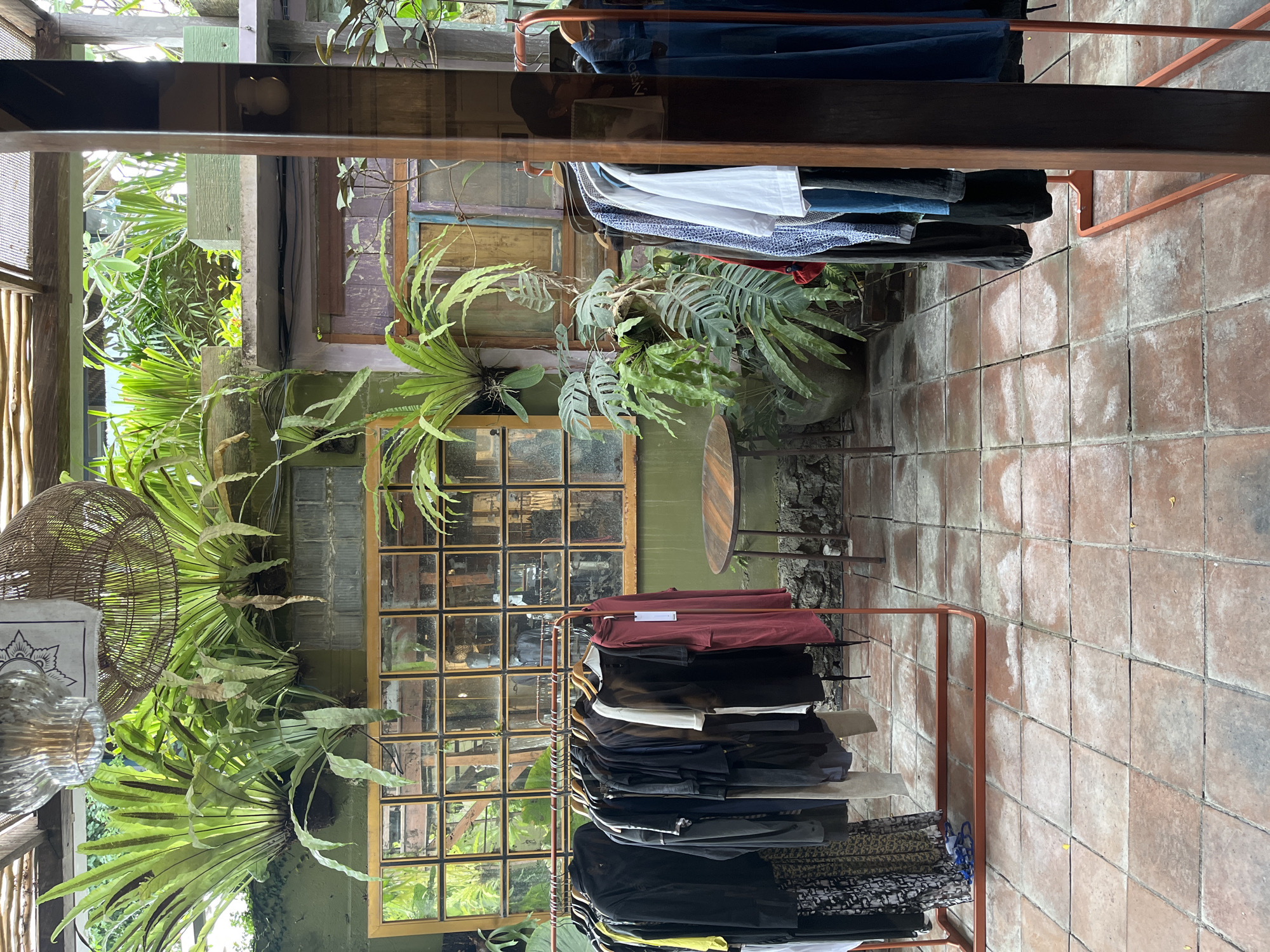 The image depicts an outdoor clothing store with a rustic, bohemian aesthetic, located in North Kuta, Indonesia. Lush greenery cascades from shelves and planters, creating a tropical atmosphere. The floor is tiled with terracotta-colored square tiles. Two clothing racks are visible; one on the left displays a variety of dark-colored garments, including dresses, shirts, and patterned skirts. The rack on the right showcases lighter-colored clothing, such as white shirts and blue denim items. In the background, a wall features a large, grid-like window with a wooden frame, reflecting the surrounding foliage. A small, round wooden table stands between the racks. The overall impression is one of a tranquil, nature-integrated retail space.