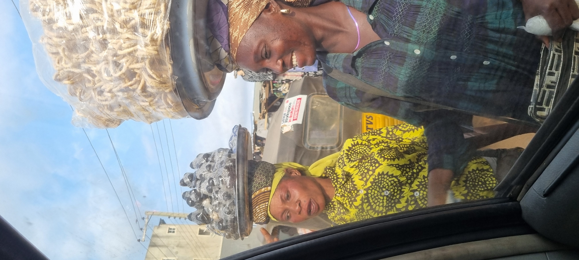 The image depicts two women selling goods on a street in Otta, Nigeria. The woman on the left wears a yellow and black patterned shirt and a yellow headscarf, balancing a tray of small, packaged items on her head. The woman on the right, wearing a green and black plaid shirt and a patterned headscarf, is smiling and looking to her right. She has a necklace and earrings and is also balancing a large tray of food items wrapped in plastic on her head. Behind them, a yellow and black three-wheeled vehicle, likely a commercial tricycle or 'keke napep', is visible, with signage that includes "TVS" and a phone number. The background suggests a busy urban environment with buildings and other market stalls, indicating a typical street scene in Otta, a city known for its industrial and commercial activities in Ogun State, Nigeria. The image appears to be taken from inside a vehicle, looking out through the window.