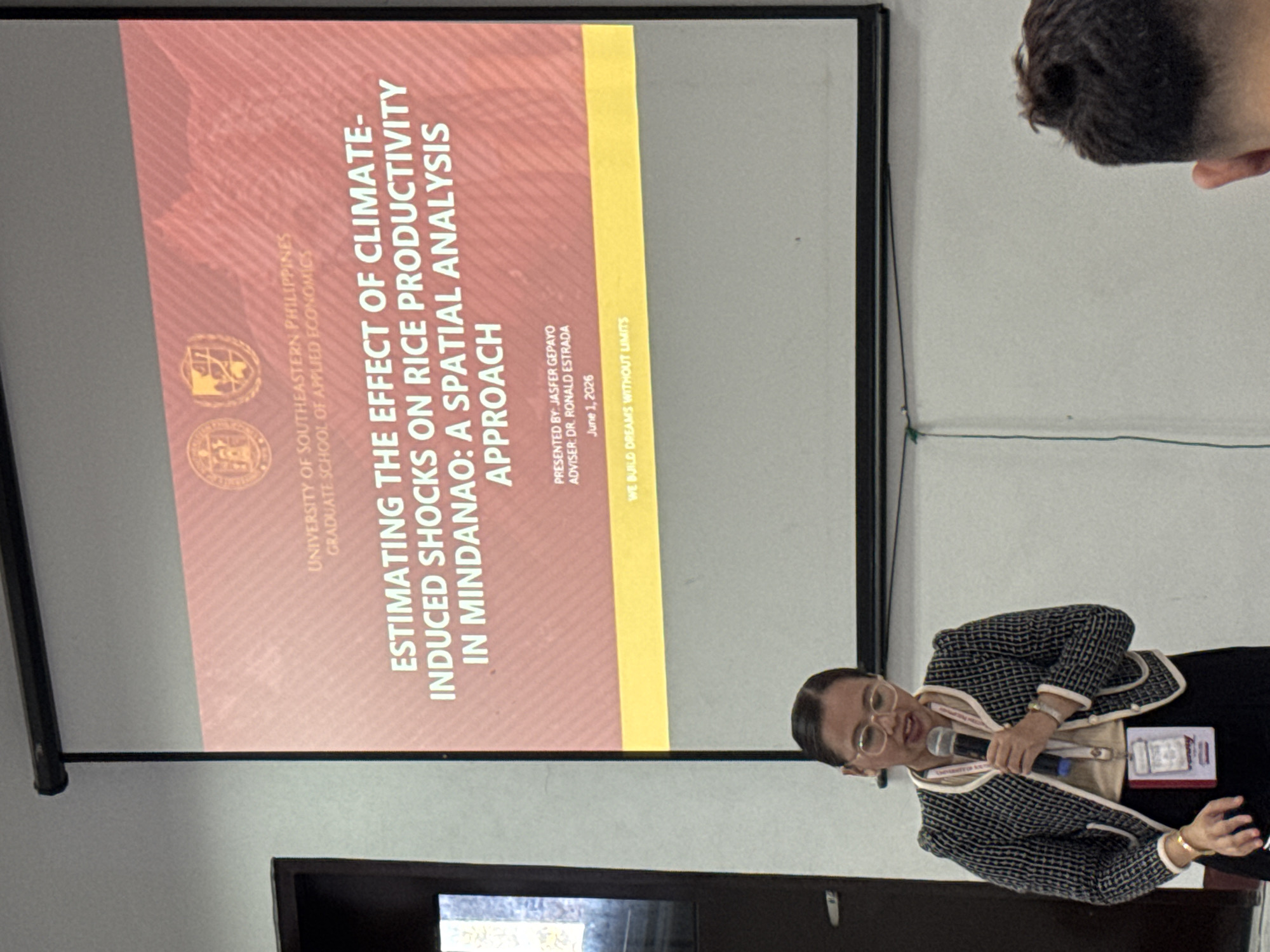 Here is a detailed description of the image:
**Scene:** The photo captures a woman giving a presentation in what appears to be a conference or seminar setting. A projector displays a slide in the background.
**People:**
- The woman in the foreground is the presenter. She wears glasses, a patterned jacket over a beige top, and a dark skirt. She is holding a microphone and gesturing with her left hand as if emphasizing a point. She has an ID badge around her neck.
- Part of a person's head is visible in the upper right corner, implying there is an audience present.
**Subjects:**
- The projector screen in the background displays the title of the presentation: "Estimating the Effect of Climate-Induced Shocks on Rice Productivity in Mindanao: A Spatial Analysis Approach." The presentation is from the University of Southeastern Philippines, Graduate School of Applied Economics. It is being presented by Jasfer Gepayo and advised by Dr. Ronald Estrada, with the date listed as June 1, 2026.
**Objects:**
- A microphone is being held by the presenter.
- An ID badge is hanging around the presenter's neck.
- A projector screen and projector are visible.
**Actions/Interactions:**
- The woman is presenting to an audience.
- She is using a microphone to speak, and gesturing with her hand.
**Environment:**
- The background appears to be a conference room or a lecture hall.