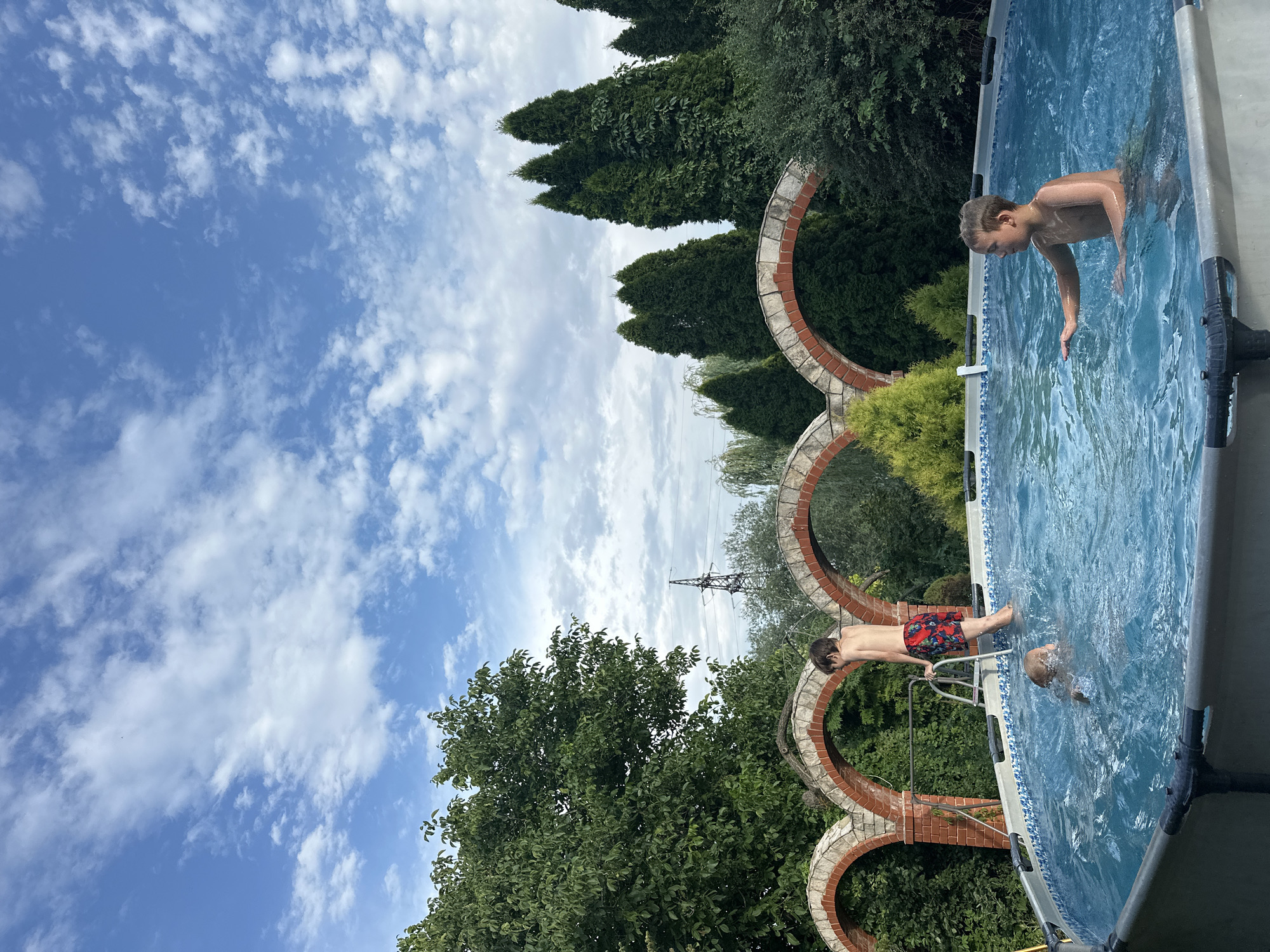 A wide-angle shot captures a summer scene in a backyard in Engels, Russia. In the foreground, an above-ground swimming pool is filled with blue water, where three young boys are playing. One boy, wearing red and black patterned swim trunks, stands on the ladder of the pool. Another boy, partially submerged, swims near the center of the pool. A third boy, with short brown hair, swims closer to the edge on the right side of the frame. The background features a brick arched structure, lush green trees, and a bright blue sky with scattered white clouds. A tall industrial tower is visible in the distance.