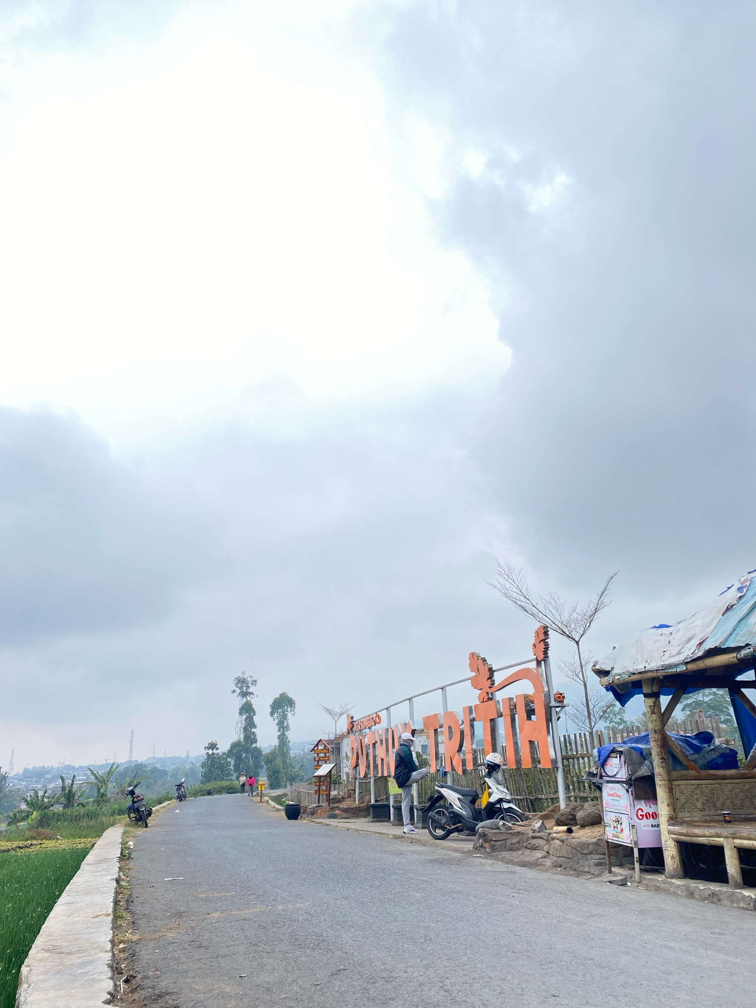 An overcast day greets the viewer at an outdoor scenic spot. In the foreground, a paved road curves into the distance, bordered by a concrete retaining wall and a field of green plants on the left. A few motorcycles are parked along the road. On the right side, a rustic wooden stall with a tarp roof offers shelter.
Dominating the middle ground is a large, orange sign with raised lettering, spelling out "TRITIH" in a stylized font. Two people stand near the sign, one leaning against a parked motorcycle. Beyond the sign, the landscape unfolds with scattered trees, buildings, and utility towers on a distant hillside. The sky is filled with heavy, grey clouds, creating a muted atmosphere.