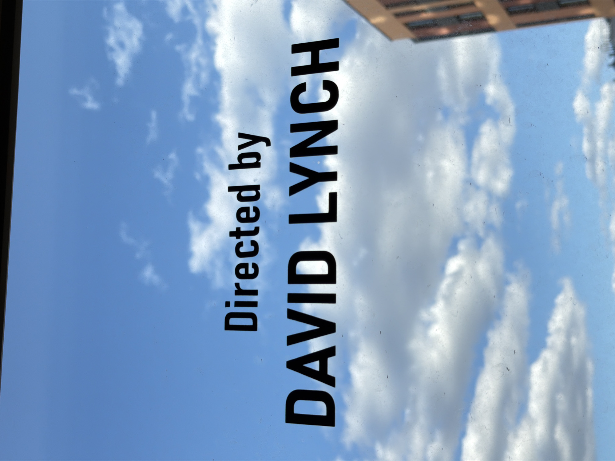 The image shows a view of the sky with clouds and a building partially visible in the top right corner. Superimposed on the sky are the words "Directed by DAVID LYNCH" in bold, black letters. There is no information to suggest this image was taken in Izhevsk, Russia.