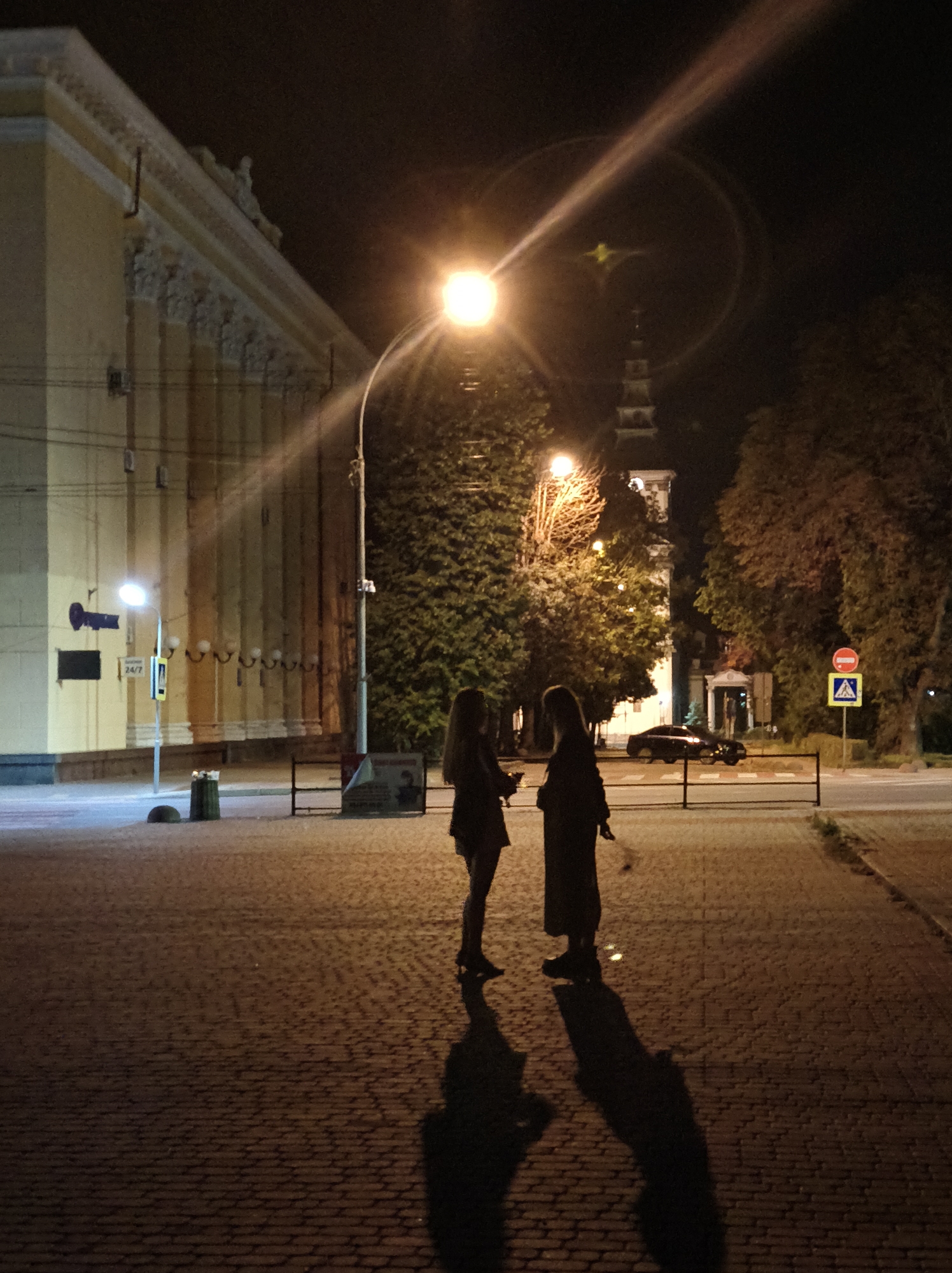 In Ternopil, Ukraine, at night, two silhouetted women stand on a cobblestone sidewalk facing each other. The woman on the left wears a short skirt and jacket, while the woman on the right is in a long dress. A streetlamp casts a bright light and lens flare above them. To the left is a building with ornate architectural details and vertical columns. A street sign with "24/7" is visible. In the background, trees and a church tower are illuminated by artificial light. A black car is parked on the street, and a pedestrian crossing is marked. Traffic signs, including a red prohibition sign and a yellow pedestrian crossing sign, are present. The ground is paved with interlocking cobblestones.