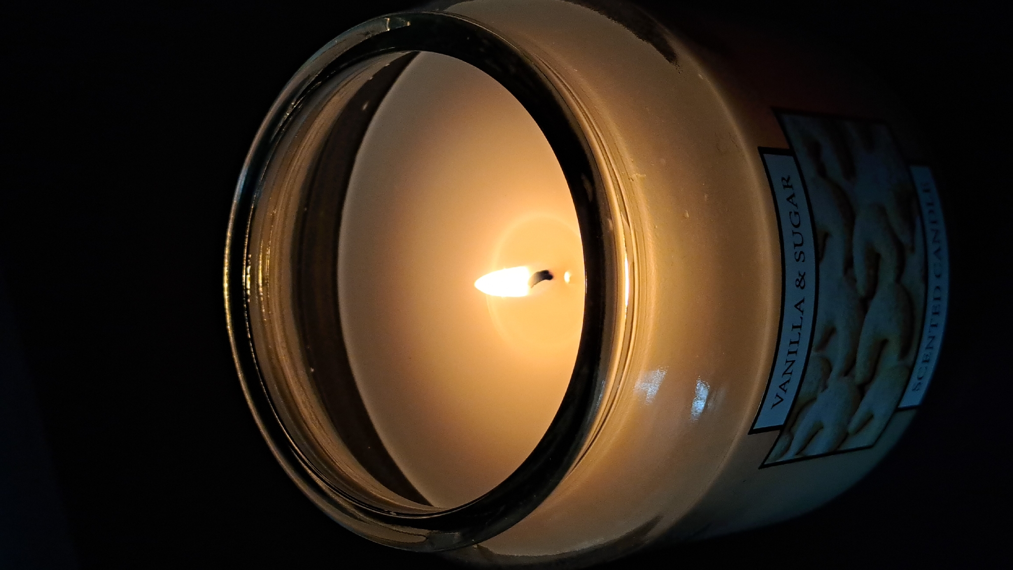 A close-up image shows a lit candle in a clear glass jar. The candle is mostly burned, revealing the flame and melted wax. A label on the jar reads "VANILLA & SUGAR SCENTED CANDLE". The background is dark, emphasizing the light from the candle. No people are present. The location in the city of Stara Zagora, Bulgaria is not identifiable from the image.