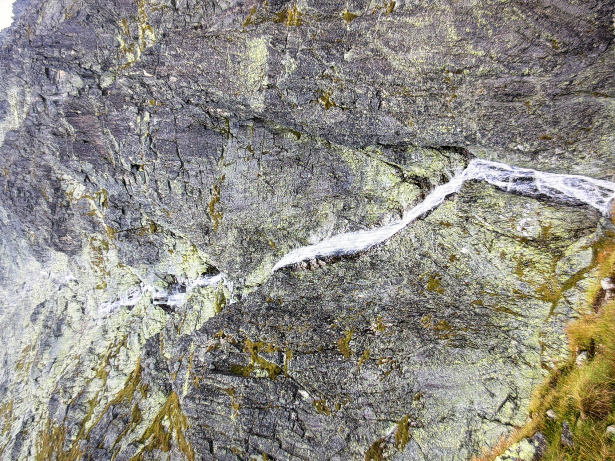 The image depicts a rocky mountainside with a waterfall cascading down. The rocks are predominantly gray and appear weathered, with patches of green moss or lichen growing on them in places. The waterfall is a narrow stream of white water flowing over the rocks. In the lower left corner, there is a patch of dry, yellowish-green grass, suggesting a natural, mountainous environment.
This image does not depict any part of the city of Kyiv, Ukraine, nor does it show any people, specific objects, or actions.