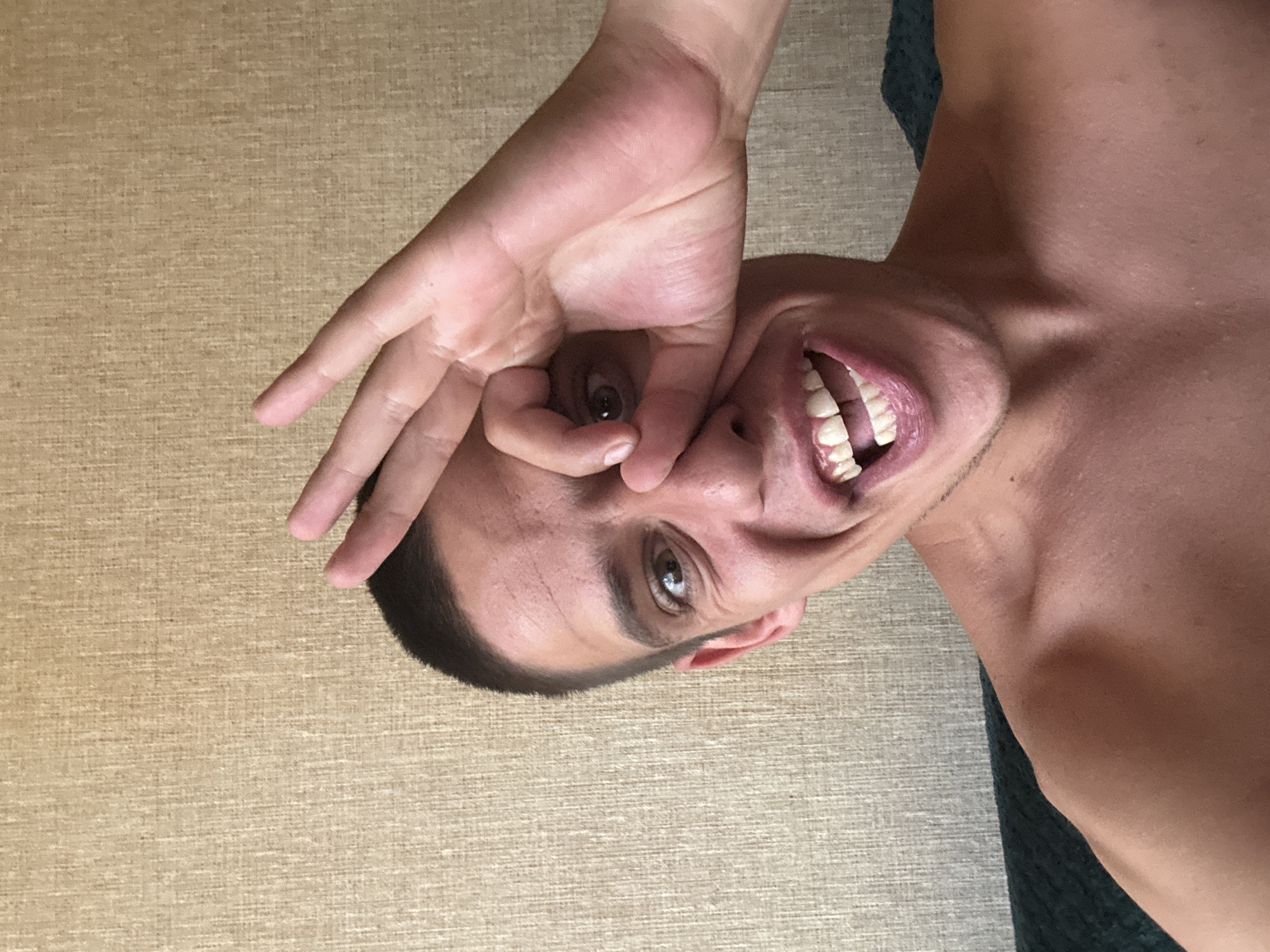 A young man, appearing to be in his early to mid-twenties, is captured in a close-up selfie. He has short, dark hair and light brown eyes. His right eye is partially obscured by his left hand, which is forming an "okay" gesture, with his thumb and index finger creating a circle. His mouth is open in a wide smile, revealing white teeth. He is shirtless, with his shoulders and upper chest visible. The background is a textured wall, possibly wallpaper, in a neutral beige or tan color. The lighting suggests an indoor environment, likely during the daytime. There is no indication of the image being taken in Sofia, Bulgaria.