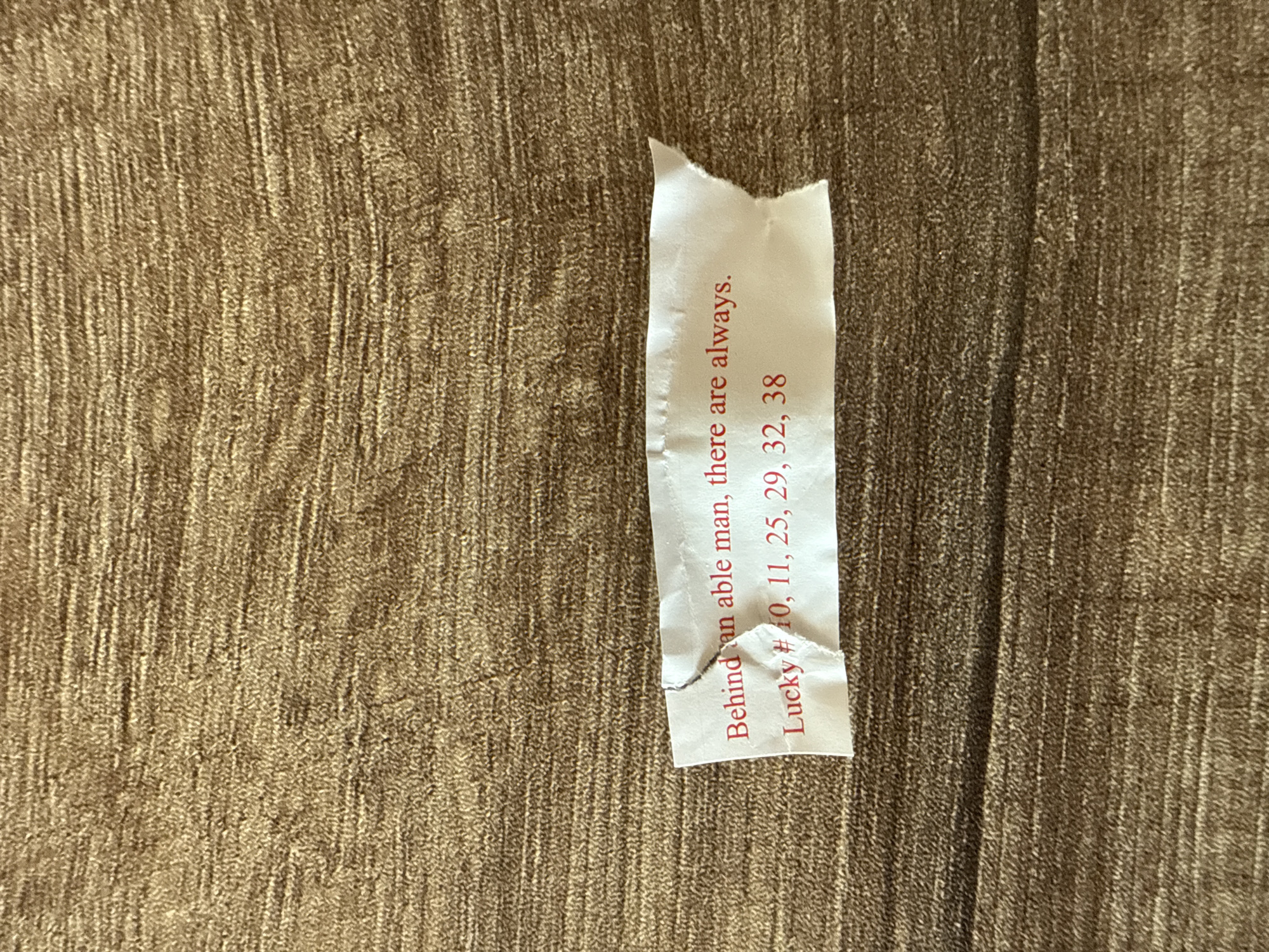 A close-up shot shows a partially torn fortune cookie slip resting on a textured, golden-brown surface. The text on the slip, printed vertically in red, reads: "Behind an able man, there are always." Below this, it states "Lucky # 10, 11, 25, 29, 32, 38." The slip is white with minor tears along the edges. No people or specific environmental elements indicating Darien, USA, are visible.