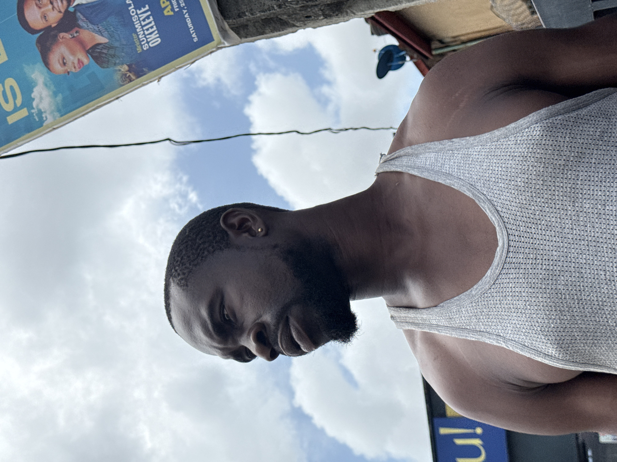 A low-angle shot shows a dark-skinned man wearing a white tank top with a beard and short hair. He is seen from the shoulders up and to the side. Behind him, there is a cloudy sky and part of a poster featuring a woman and a man, with text that includes "Minister SUNMISOLA OKELEYE" and "SATURDAY, 25". A blue sign with a yellow "u" is also visible. The location appears to be a street in Ado, Nigeria.