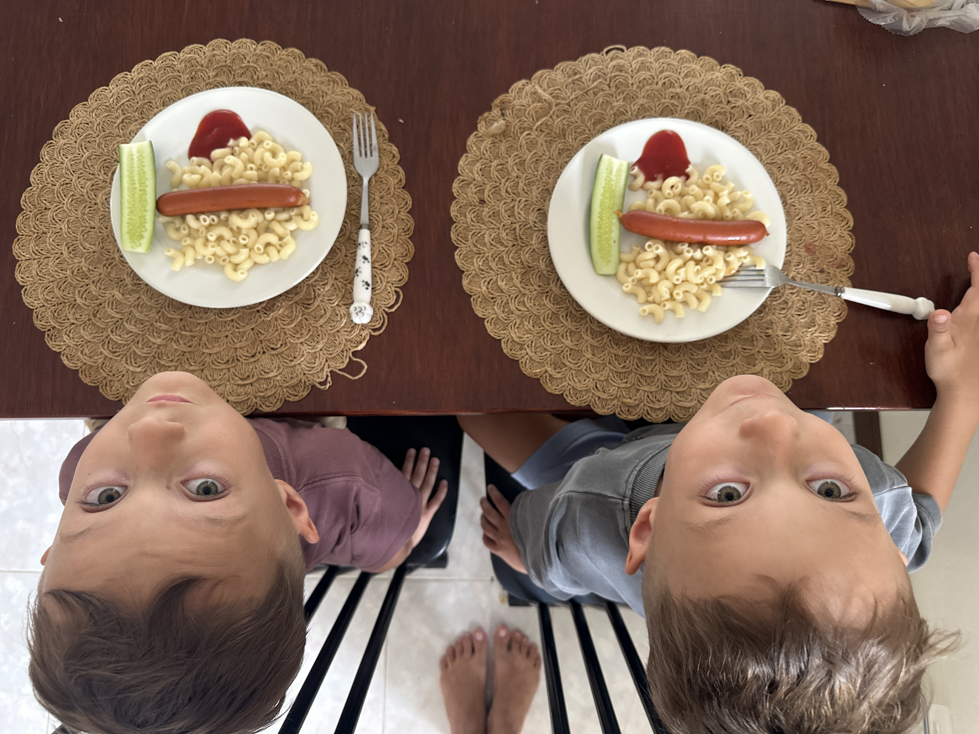 Two young boys are seated at a wooden table, looking up at the camera, which is positioned above them. They are each served a plate of macaroni pasta with a hot dog, a slice of cucumber, and a dollop of ketchup. The plates are placed on circular woven placemats. Forks with patterned handles are on the right side of each plate, with one fork partially visible in the hand of the boy on the right. The boys are wearing casual clothing, one a maroon long-sleeved shirt and the other a grey t-shirt. Their expressions are curious and engaged. The scene appears to be an indoor setting, possibly a home or dining area, given the table and chairs. The lighting suggests it is daytime.