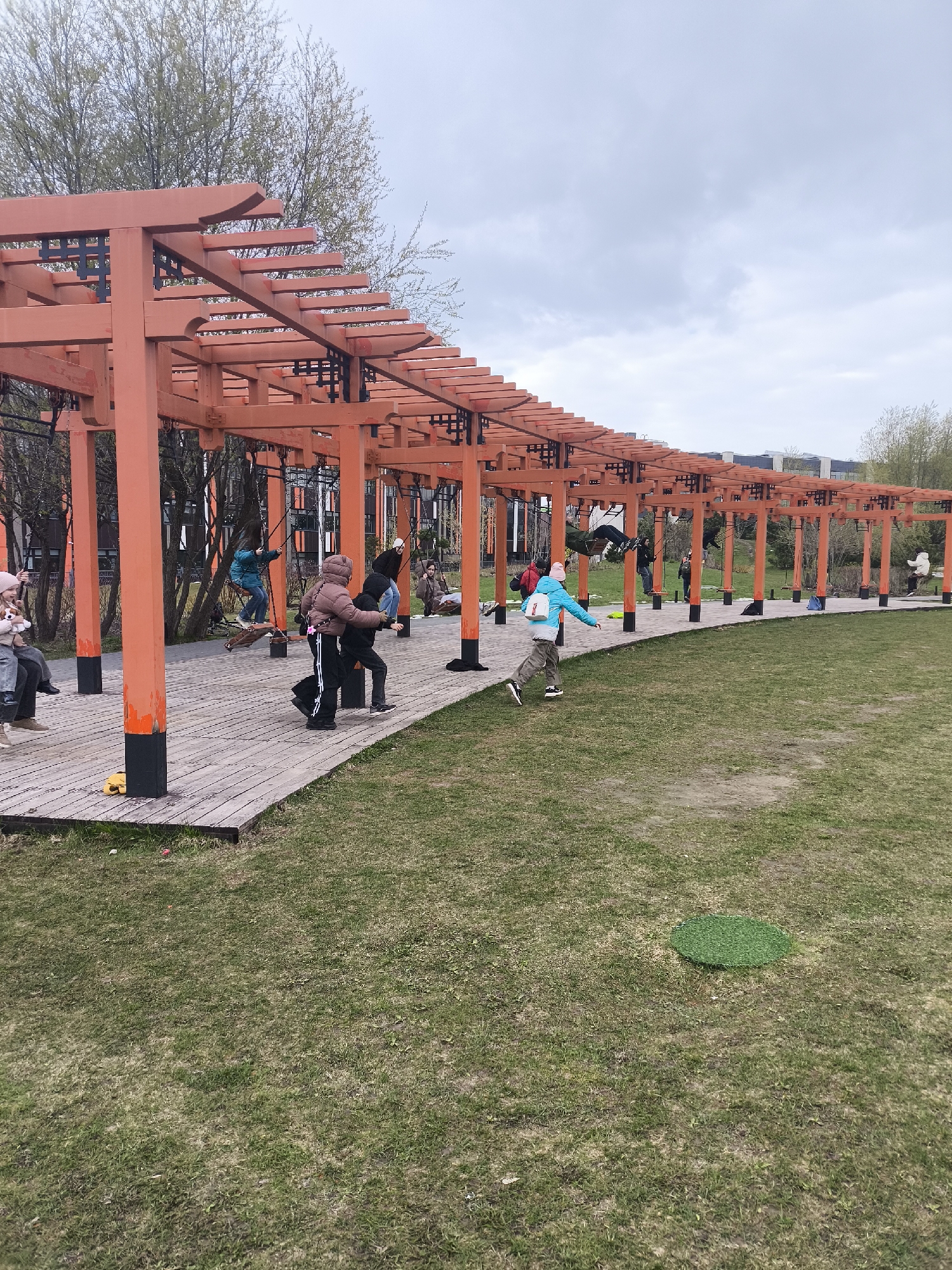 This media file captures an outdoor recreational area, likely a park, in Moscow, Russia, featuring a distinctive orange and black wooden structure resembling a pergola or a series of interconnected torii gates. The structure forms a long, curving arcade over a wooden boardwalk pathway.
Several people, predominantly children and young adults, are present and engaged in activities. Many are utilizing the swings suspended from the structure's crossbeams. One child in a blue jacket and white backpack is running on the wooden path, seemingly in motion. Another person in a brown hooded jacket appears to be walking alongside someone in a black outfit. Other individuals are visible in the background, either swinging or standing on the pathway.
The scene is set under an overcast sky, suggesting a cloudy day. The time of day appears to be daytime. Many individuals are dressed in warm clothing, including jackets and hats, indicating cool weather. The ground to the right of the wooden pathway is covered in green grass, with some patches appearing drier or sparser. A small, circular patch of bright green artificial turf is visible on the grass in the foreground. The wooden structure itself has prominent orange pillars and beams with black accents and intricate joinery. In the background, deciduous trees with budding leaves and modern buildings can be seen. There is no visible text in the image.