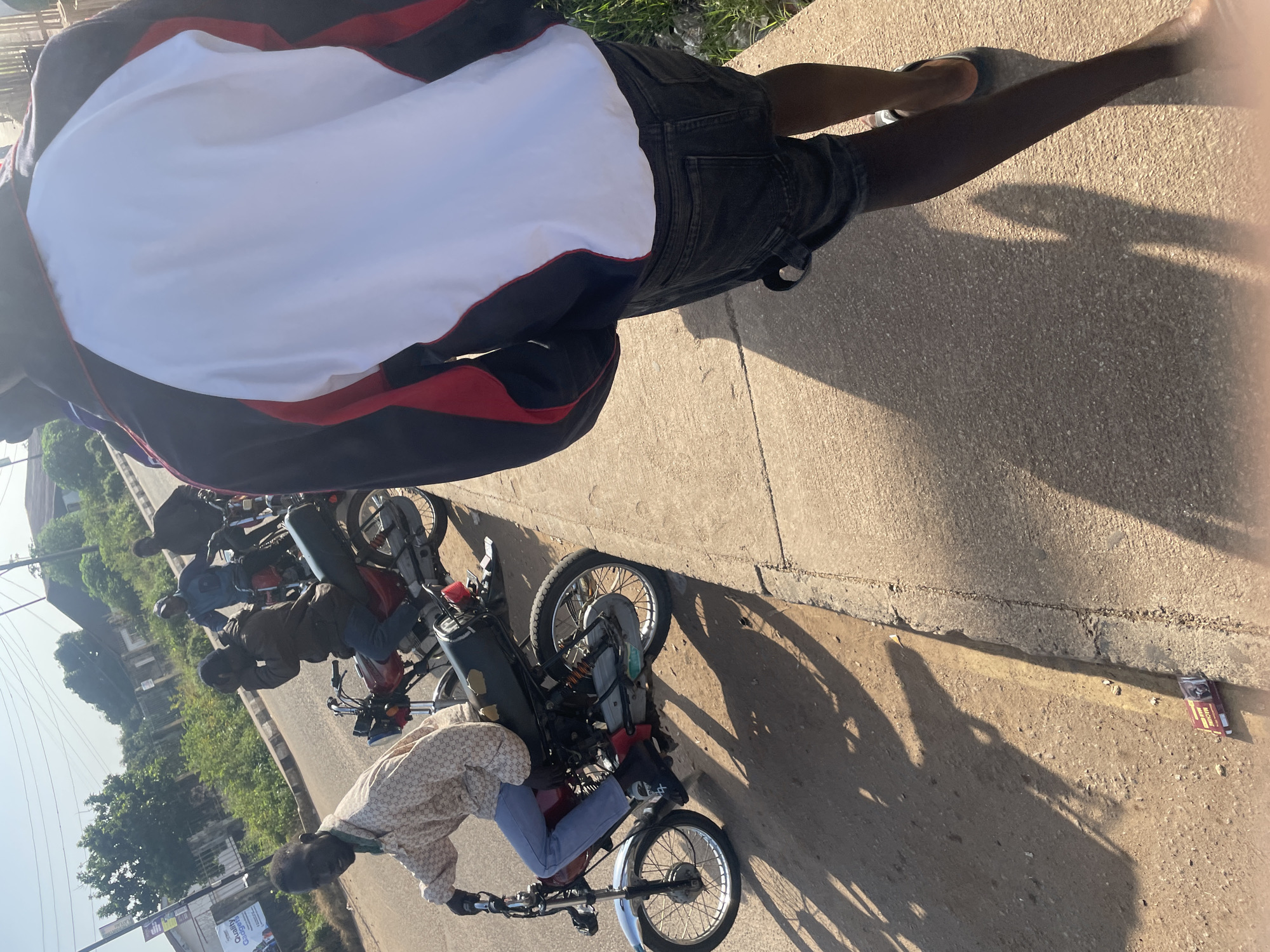 In Jos, Nigeria, on a sunny day, three motorcycles are parked on the side of a road. The foreground features a person walking away from the camera, wearing a jacket with white and red accents and dark denim shorts. Behind this person, a man on a motorcycle is looking towards the camera, dressed in a patterned shirt and blue jeans. He is wearing red shoes. Another motorcycle is positioned behind him, with two individuals seated on it. The road is bordered by a concrete sidewalk and grassy verges. Sunlight casts long shadows across the scene, indicating it is likely late afternoon. A crumpled pack of cigarettes lies on the ground near the sidewalk.