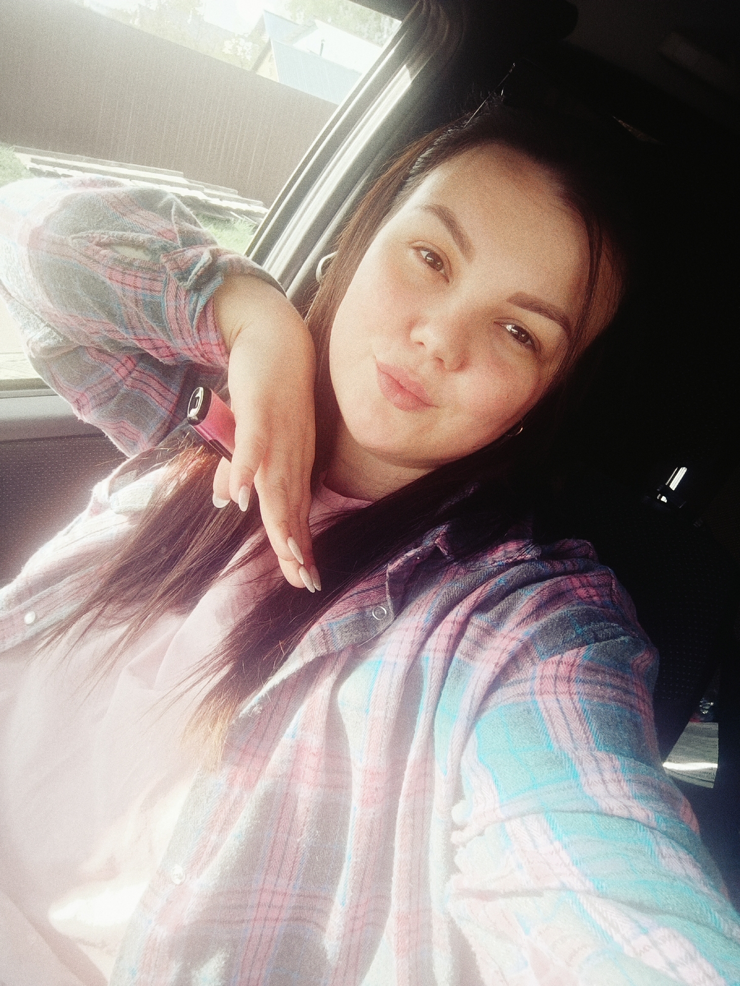 A young woman is sitting in a car, likely in the city of Chaikovsky, Russia, though there is no specific identifying detail in the image to definitively determine that location. She is turned slightly towards the camera, her face in focus. She has dark hair and is wearing a pink t-shirt covered by a pastel plaid shirt. Her left hand is raised to her face, holding a pink electronic cigarette. The car interior is dark, with the window and outdoor scenery partially visible in the background. The lighting is bright and sunny.