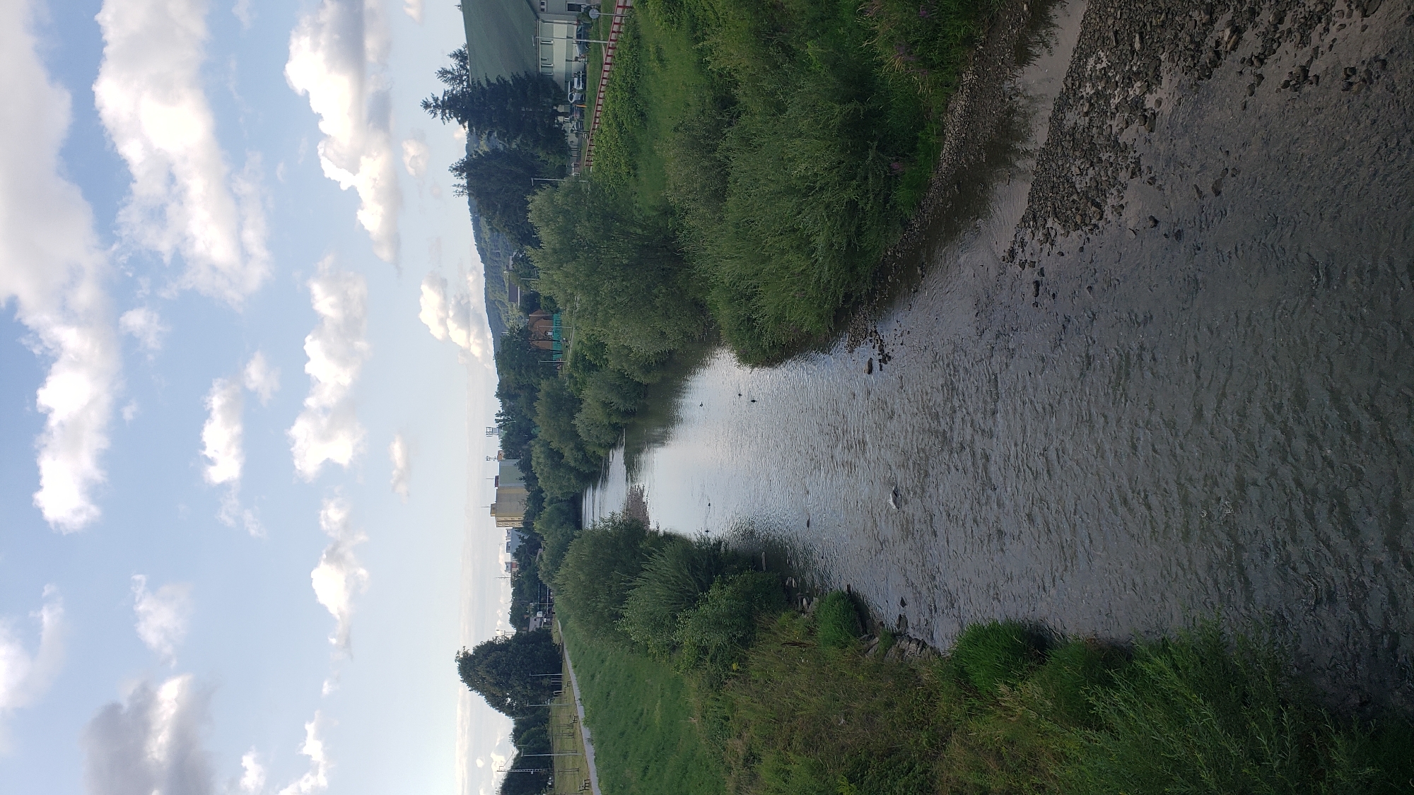 The image shows a view from a bridge looking down at a river flowing through a city. The river is bordered by lush green vegetation on both sides. The left bank of the river features a paved walkway lined with trees and benches, suggesting a park or recreational area. In the distance, buildings and structures of the city of Prešov, Slovakia, are visible, nestled against a backdrop of rolling hills. The sky is partly cloudy, with fluffy white clouds scattered across a pale blue expanse. The overall scene depicts a tranquil urban landscape where nature and city life coexist.