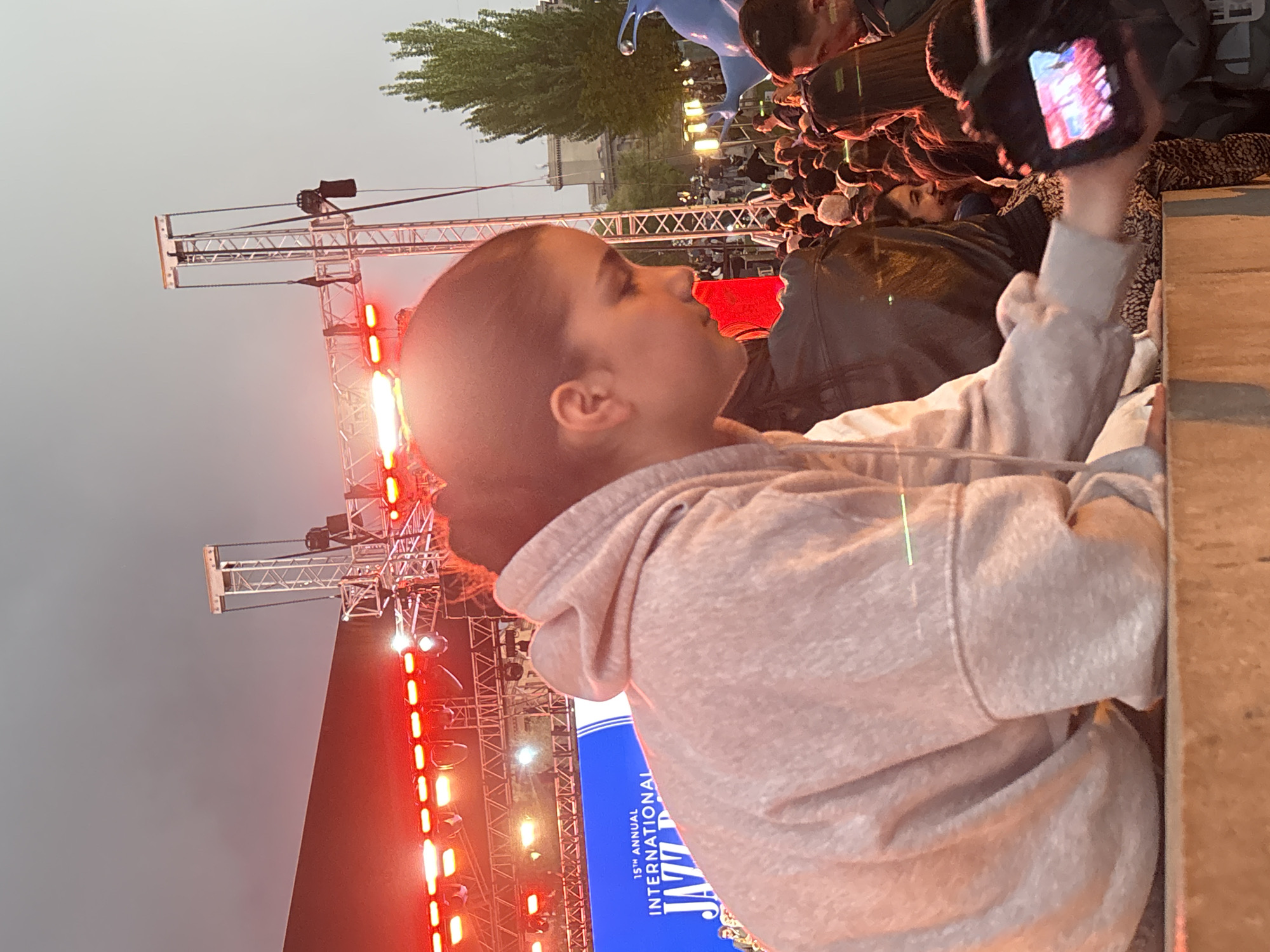 The image captures an outdoor evening event in Yerevan, Armenia, likely a concert or festival. The primary subject is a young person, seen in profile on the left, looking upwards and to the left with a focused expression. They have short dark hair and are wearing a light-colored, possibly grey, hoodie. In the foreground on the right, a wooden-like barrier or ledge is visible.
In the background, a stage setup is prominent, featuring tall metal truss structures adorned with numerous bright red and orange lights, illuminating the scene. Behind the main subject and extending into the mid-ground, a dense crowd of people is gathered, suggesting a well-attended event. Some individuals in the crowd are partially visible, including one person holding up a smartphone, seemingly recording the event.
To the left of the main subject, part of a vertical banner is visible against the stage lights. The text on the banner reads "15th ANNUAL INTERNATIONAL JAZZ D", indicating it's related to a jazz event, likely a "Jazz Days" or "Jazz Festival". Further in the background, trees are visible against the dark sky, and a large blue sculpture or object can be faintly seen above the crowd. The overall atmosphere is lively, characteristic of an evening public performance.