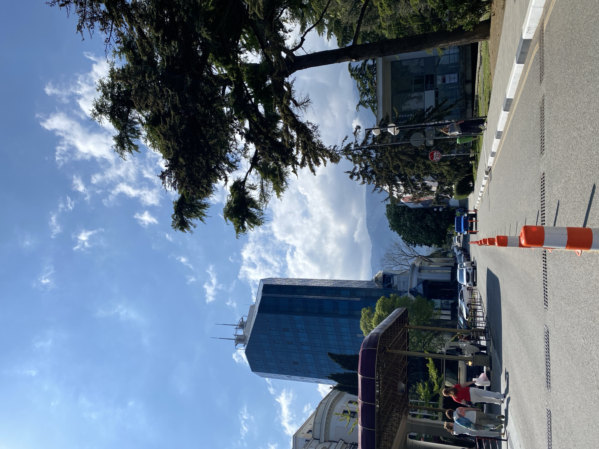 The image shows a street scene in the city of Ялта, Ukraine. The perspective is from a low angle, looking upwards towards a tall, modern blue glass building. The sky is visible, with a mix of blue and white clouds. In the foreground, on the right side of the frame, is a street lined with orange and white traffic cones. A row of parked cars is visible along the side of the street opposite the cones. People are standing on the sidewalk near a structure with a dark purple roof. A tree is prominent on the left side of the frame, partially obscuring the view of the building. In the background, a mountain range can be seen partially obscured by clouds.