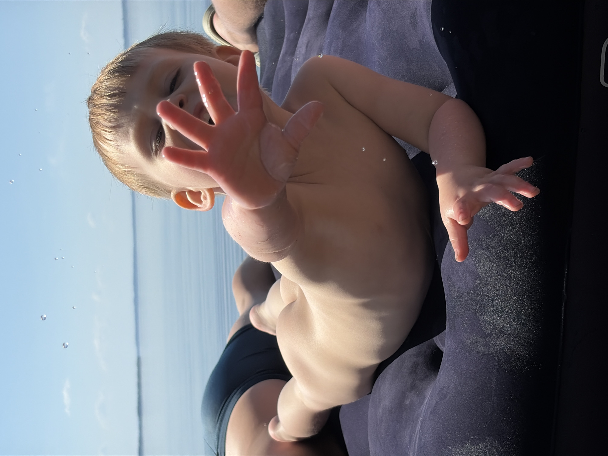 A child lies on an inflatable raft. The child's right hand is extended towards the viewer. Water droplets cling to the child's hand and skin. A person is partially visible behind the child, wearing dark-colored swimwear. The background shows a body of water and a clear blue sky. The scene is bathed in bright sunlight. The geographic location is Zabegaevo, Russia.