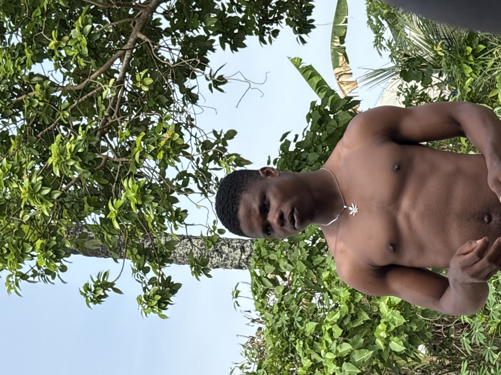 A medium shot captures a shirtless, dark-skinned man standing in a lush, green outdoor environment. The man is facing slightly to the left, with a silver necklace around his neck. The background features a variety of trees and foliage, including broad leaves and banana trees, suggesting a tropical climate. The scene may be located in the city of Ikot Ekpene, Nigeria based on the location, based on the presence of the flora, the presence of the African man, and the cultural cues within the image.