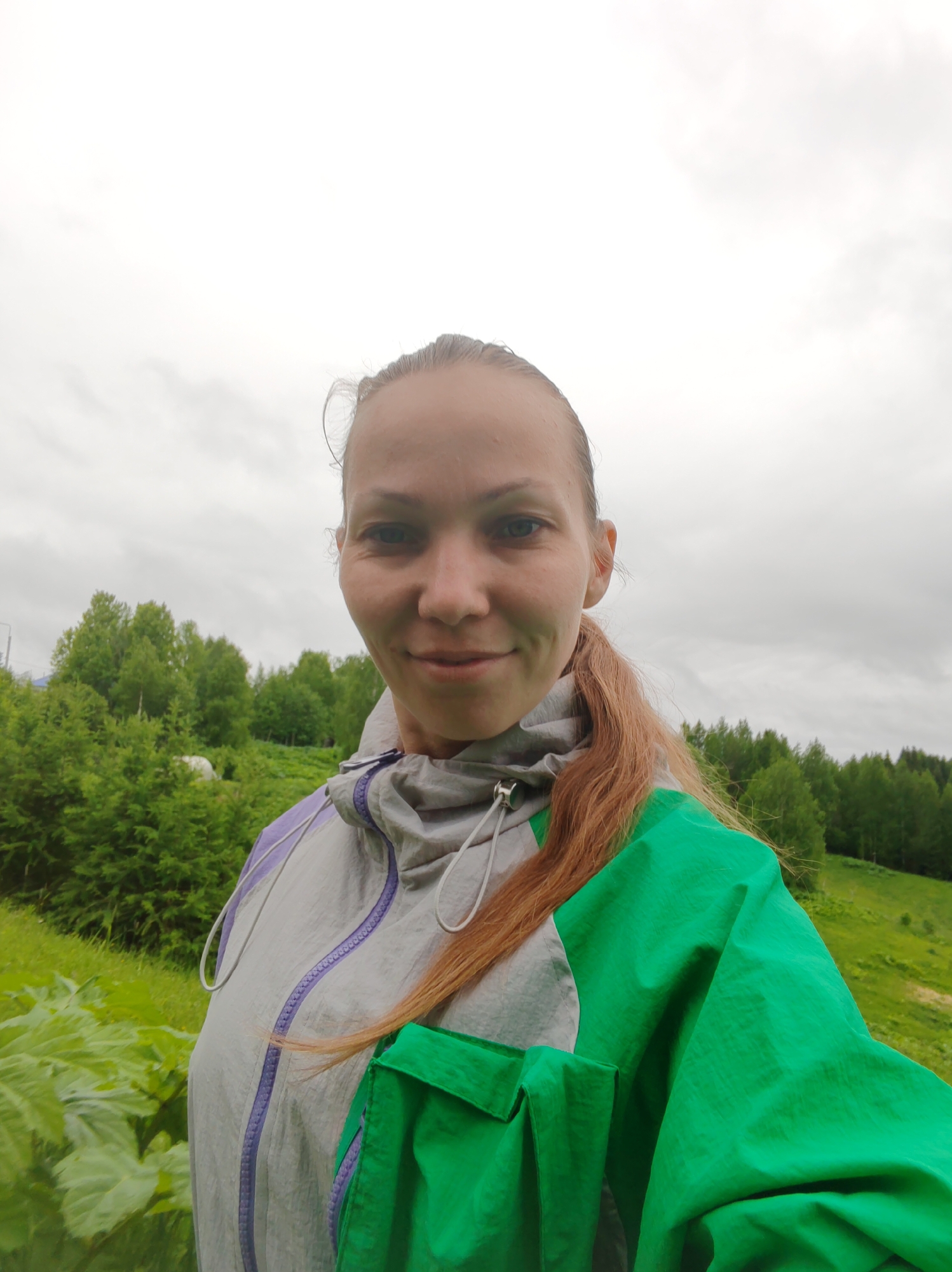 A woman is captured in a selfie outdoors in Чухлэм, Russia. She wears her reddish-brown hair tied back in a ponytail that rests over her right shoulder. She is dressed in a multi-colored windbreaker jacket, featuring grey on the left side, bright green on the right, and subtle purple zipper and trim details. A gentle, faint smile is present on her face as she looks directly at the camera.
The setting is a lush, natural landscape under an overcast sky, indicating cloudy daytime weather. In the background, a dense forest of various green trees covers rolling hills. Large, vibrant green leaves are visible in the immediate foreground, suggesting abundant plant life. The overall scene conveys a casual moment in a serene, natural environment. No text is visible in the image.
