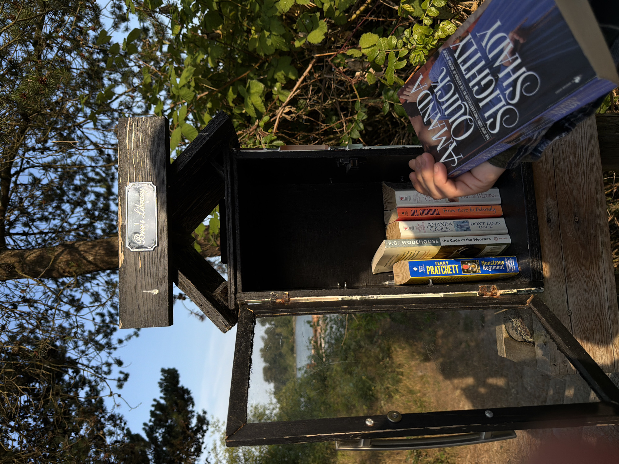 A person's hand holds an open book titled "Slightly Shady" by Amanda Quick. The book is held in front of a small, open wooden structure containing several other books. The structure is painted black and appears to be a "little free library". The background shows trees, foliage, and a body of water. The location is identified as a little free library at the Port Coquitlam Public Library in Port Coquitlam, Canada. The lighting suggests it is daytime, with sunlight illuminating the scene. The overall atmosphere is natural and serene.
