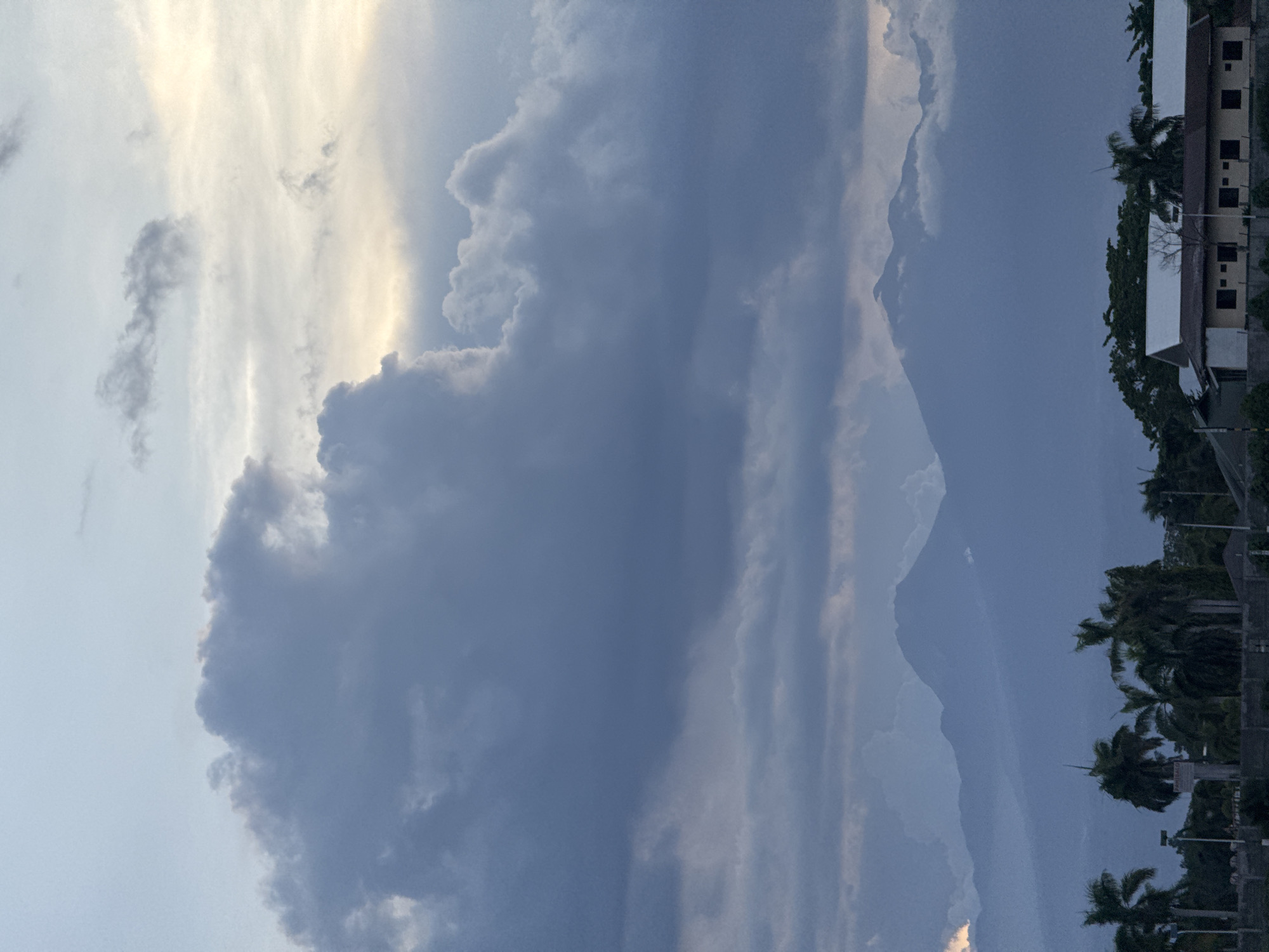 The image depicts a sky filled with dramatic, layered cloud formations in Davao City, Philippines. The clouds range in color from bright white and pale yellow where the sun shines through to dark gray and blue. In the distance, a hazy mountain range is visible under the cloud cover. Along the right edge of the image, buildings and palm trees are seen, suggesting an urban or suburban setting. The image has a slightly rotated orientation.