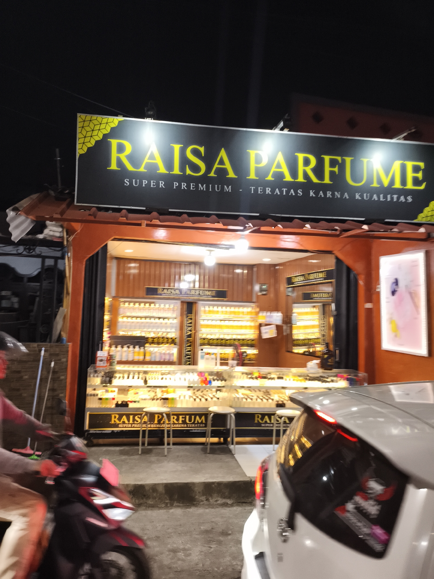 In the city of Depok, Indonesia, at night, a street-level view shows a storefront illuminated by artificial light. The storefront features a prominent black sign with yellow lettering that reads "RAISA PARFUME" and below it, "SUPER PREMIUM - TERATAS KARNA KUALITAS." Inside the store, rows of perfume bottles are displayed on shelves. To the left of the storefront, a person on a motorcycle is partially visible, captured in motion. In front of the store, a white car is parked, its rear visible. The scene is characteristic of a small business operating in an urban environment in Depok.