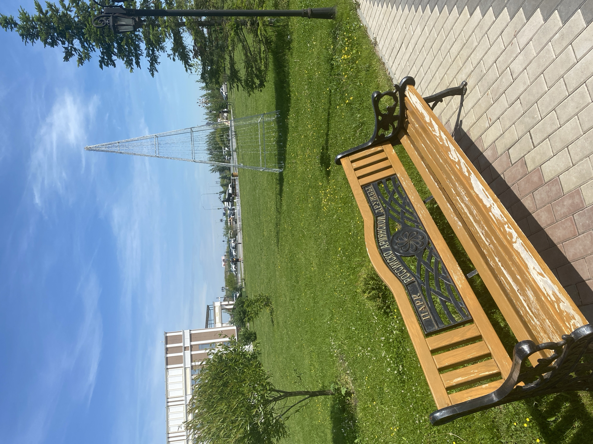 This image captures a bright, sunny scene in an outdoor park or green space. The foreground is dominated by a wooden park bench with black, ornate cast-iron arms and backrest frame. The wooden slats of the bench, particularly on the seat, show significant wear and peeling light-colored paint, revealing the natural wood underneath.
On the backrest of the bench, a metal plaque is integrated into the cast-iron frame, featuring Cyrillic text. The text reads: "ПАРК РОССИЙСКО-АРМЯНСКОЙ ДРУЖБЫ", which translates to "PARK OF RUSSIAN-ARMENIAN FRIENDSHIP".
To the right of the bench, a paved pathway made of interlocking light-colored bricks (beige/light grey and some reddish-brown hues) runs alongside a strip of green grass. The grass area extends into the middle ground and background, dotted with small yellow flowers.
In the middle ground, a large, conical metal framework structure stands tall, possibly a decorative installation or a frame for a large holiday tree. Further back on the left, a modern building with brown and white facades is visible. Small trees with green foliage are scattered across the grassy area.
The sky is clear and bright blue with a few wispy white clouds, indicating fair weather. Strong sunlight casts distinct shadows, suggesting it is midday or early afternoon. A black lamppost with an ornate design is visible at the upper left, partially obscured by the branches of a coniferous tree (likely a pine or fir). Several parked cars can be seen in the far distance along a road in the background. No people are visible in the scene.