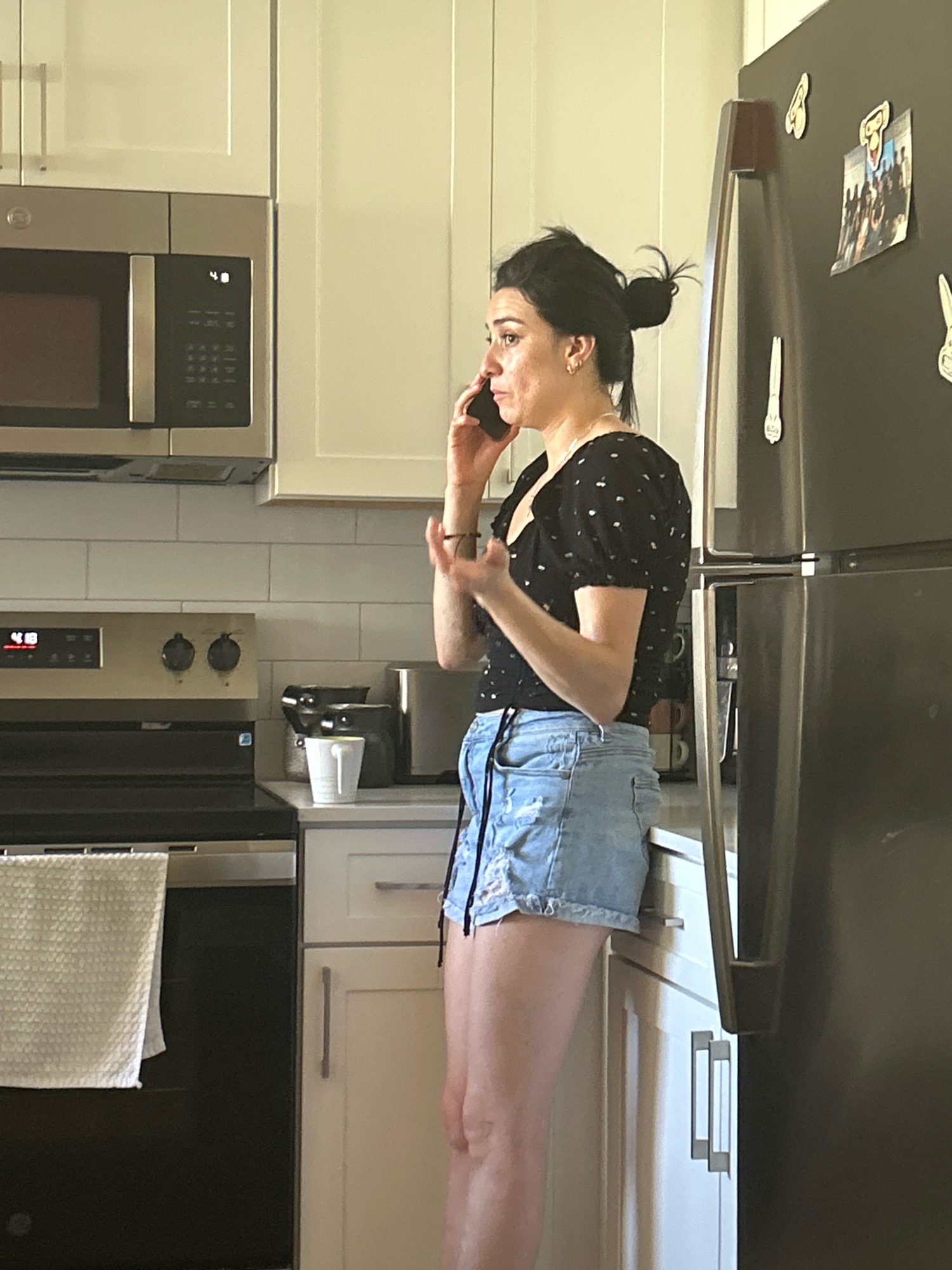 A woman with dark hair pulled into a bun is standing in a kitchen and talking on a cell phone. She is wearing a black top with white polka dots and light blue denim shorts. She has one hand up, gesturing as she speaks on the phone. The kitchen has white cabinets, a stainless steel microwave above a stovetop, and a large stainless steel refrigerator with magnets on the door. A white towel is hanging on the oven. The scene is indoors. The city of Lake Forest is in the United States of America.