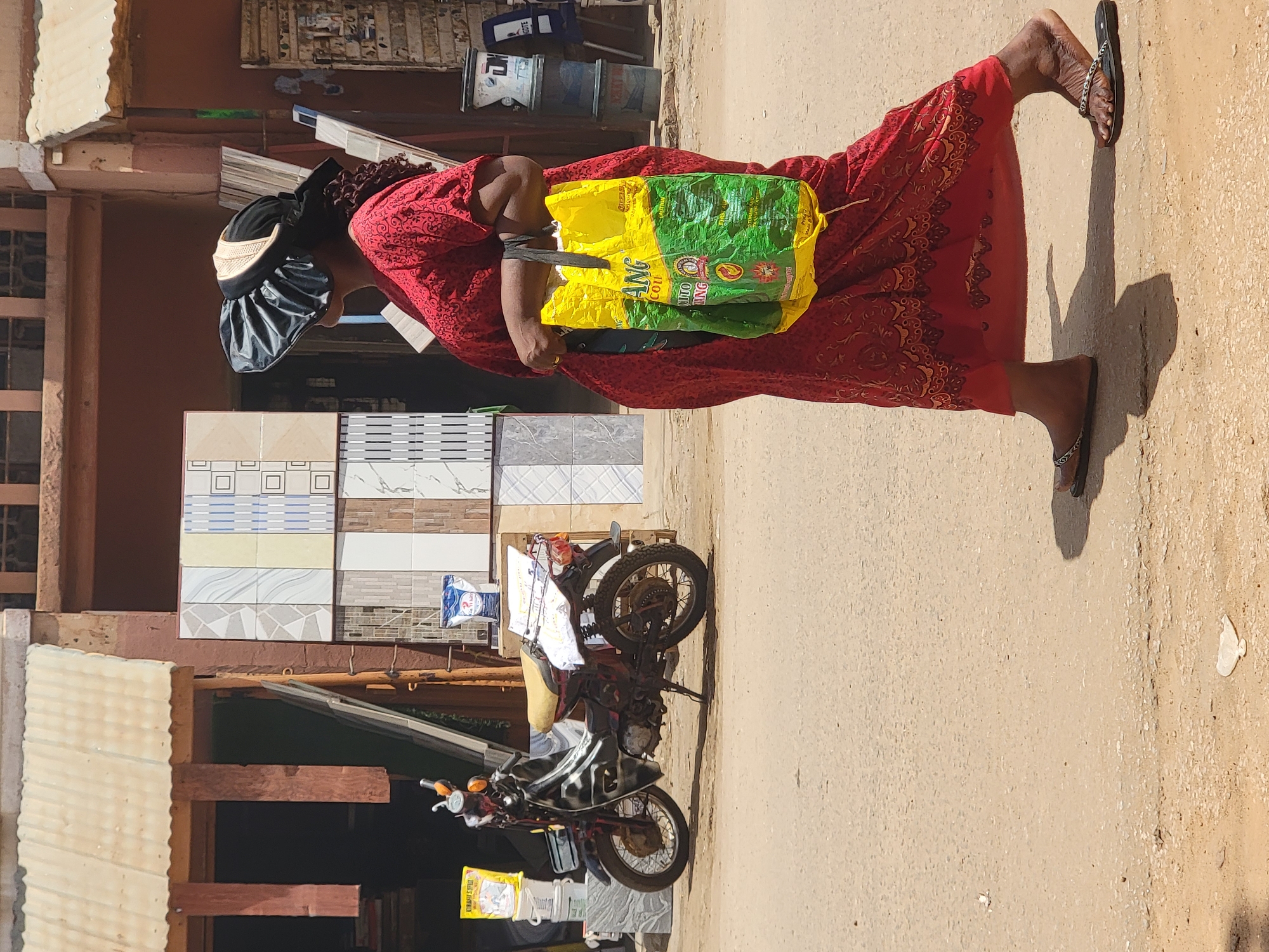 A woman in a red dress and a satin hair bonnet walks on a dusty road in Maiburiji, Nigeria. She carries a large, brightly colored bag (yellow and green) attached to her arm with a black tie. The road is unpaved and has a light brown/tan color. To the left of the road is a building with tile samples displayed outside. A parked motorcycle laden with bags stands near the building. The lighting indicates a sunny day.