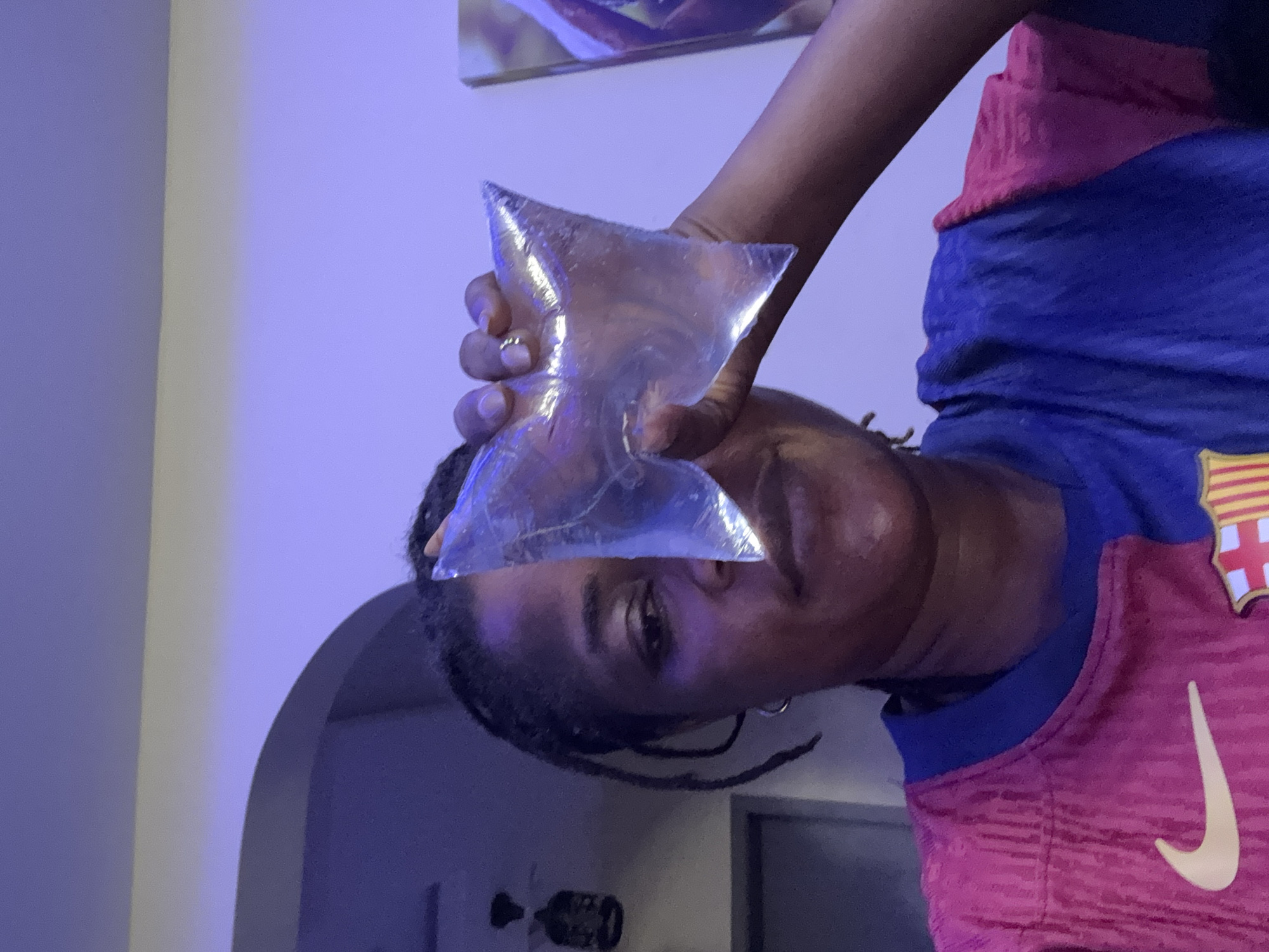 The image is a close-up, low-angle shot of a Black woman holding a clear plastic bag filled with liquid in front of her face, obscuring her right eye. She is looking towards the camera with her left eye visible. She has braided hair and is wearing a brightly colored shirt, half pink and half blue, with a Nike logo and what appears to be a FC Barcelona crest. The lighting in the image is predominantly purple. The background is dimly lit, showing a portion of a ceiling and a wall. There is no discernible information in the image to indicate a specific location in Ajara, Nigeria.