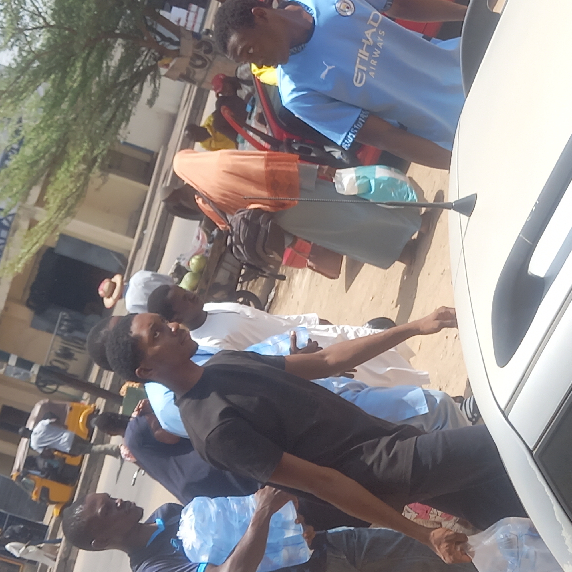 Dadi Riba, Nigeria: A group of people are seen on a sandy street beside a white car. One man in a blue Manchester City jersey stands near the car, while another man in a black t-shirt stands with his arm outstretched. A woman wearing an orange hijab stands in the background. Other individuals are carrying plastic bags filled with items. A yellow vehicle is parked on the left side of the frame. A tree is partially visible in the upper left corner. Buildings line the street.